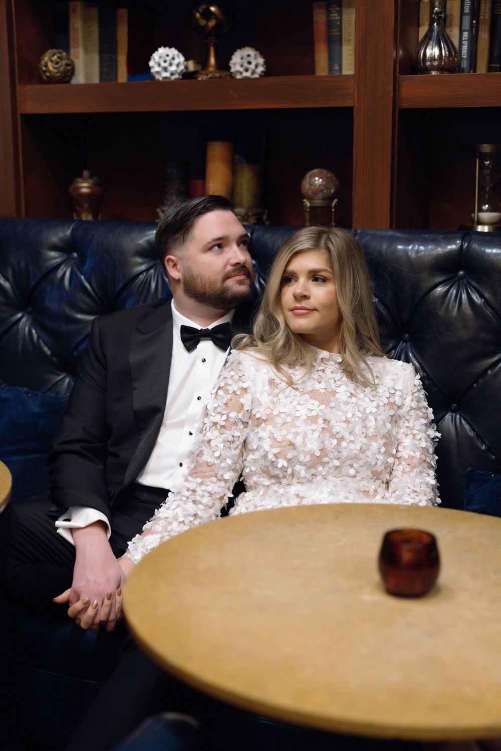 A couple in formal attire sits at a round wooden table in a room with dark leather seating and bookshelves.