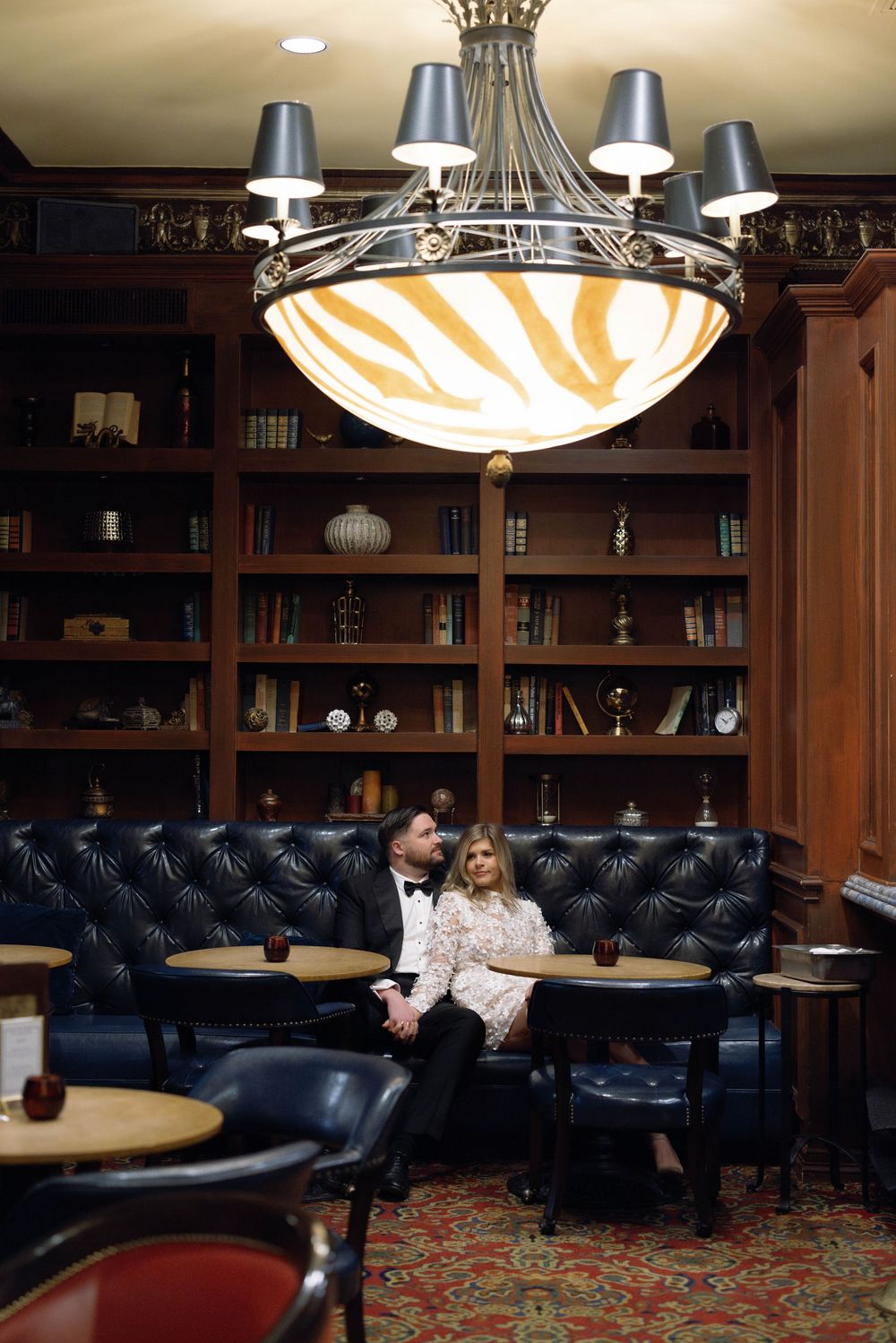 Elegant library lounge with leather banquettes, wooden bookshelves and ornate ceiling chandelier in warm ambient lighting.