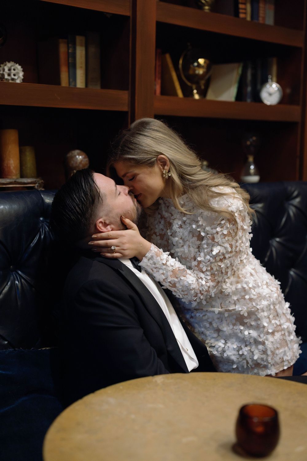 A romantic moment between newlyweds sharing a kiss at a table with a detailed lace wedding dress visible.