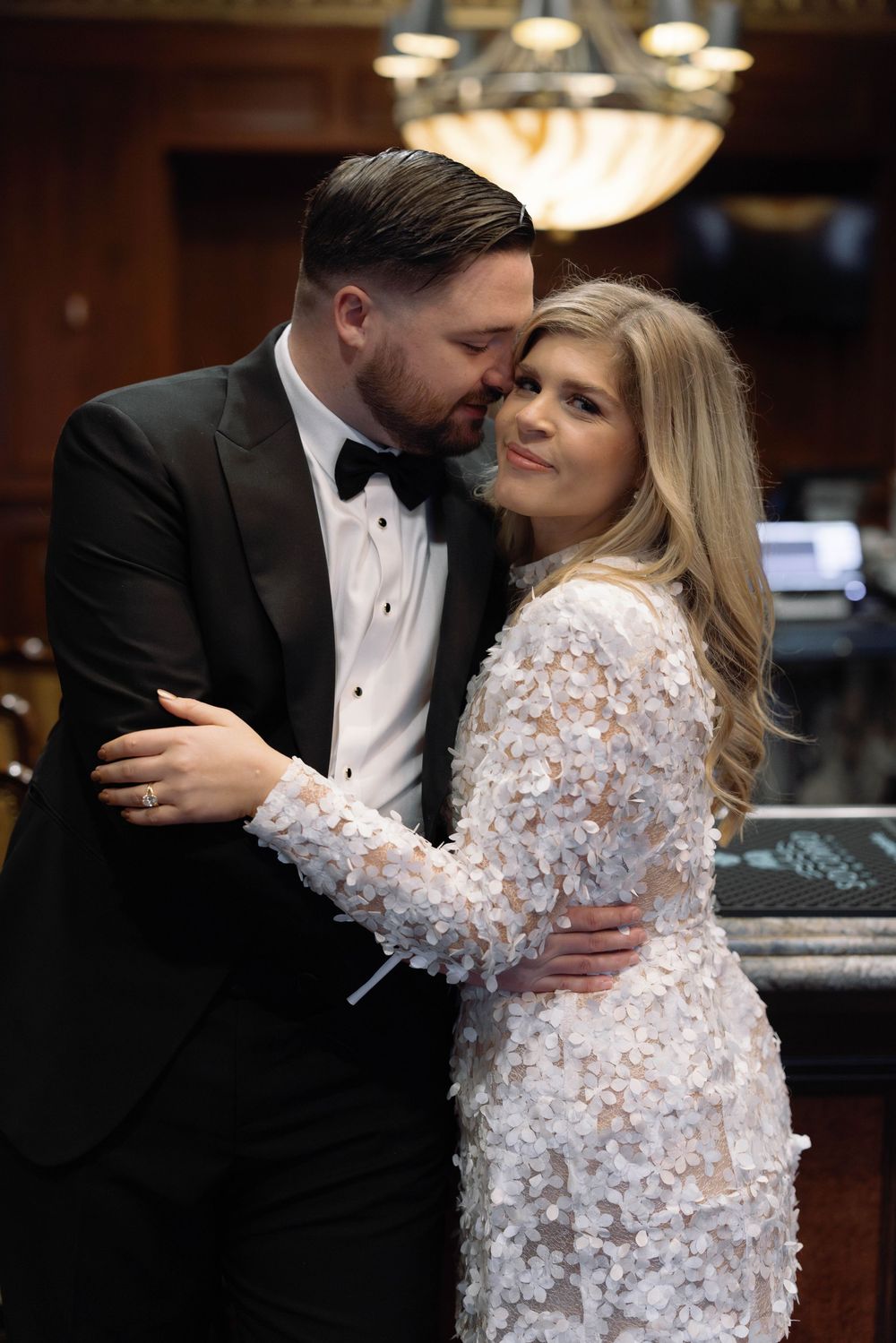 A bride in a white beaded gown and groom in black tuxedo embrace intimately at an elegant indoor venue.