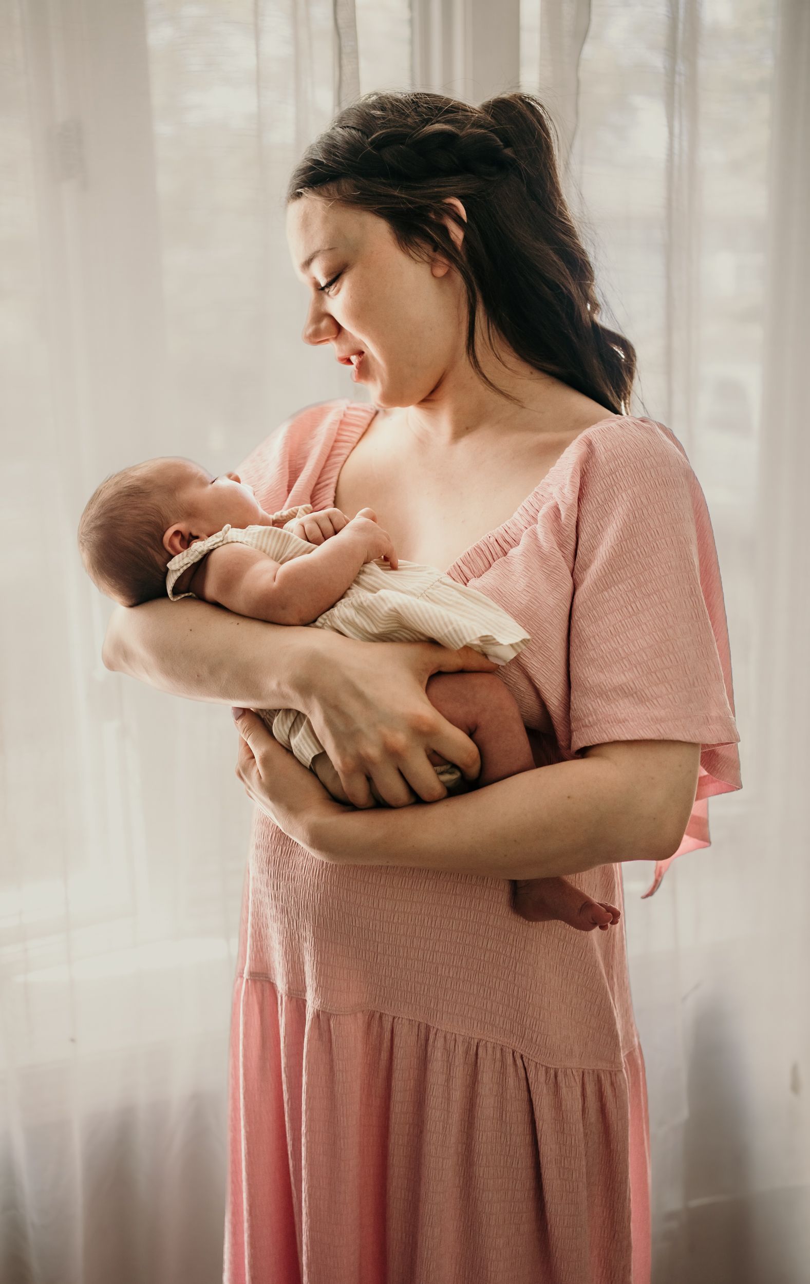 Mother in a pink dress gazes lovingly at her newborn baby near a sunlit window.