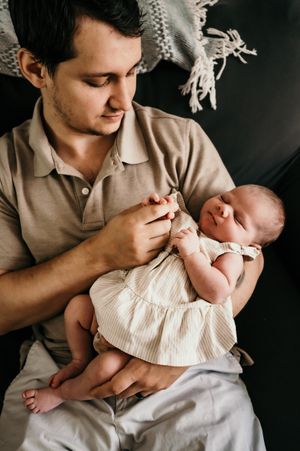 Parent in beige polo shirt tenderly holds sleeping newborn wearing white dress.
