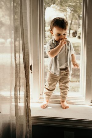 Toddler stands on windowsill in casual outfit with thoughtful expression.