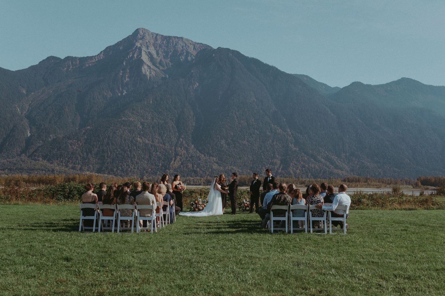 An outdoor wedding ceremony takes place in a grassy field with dramatic mountain peaks rising in the background.