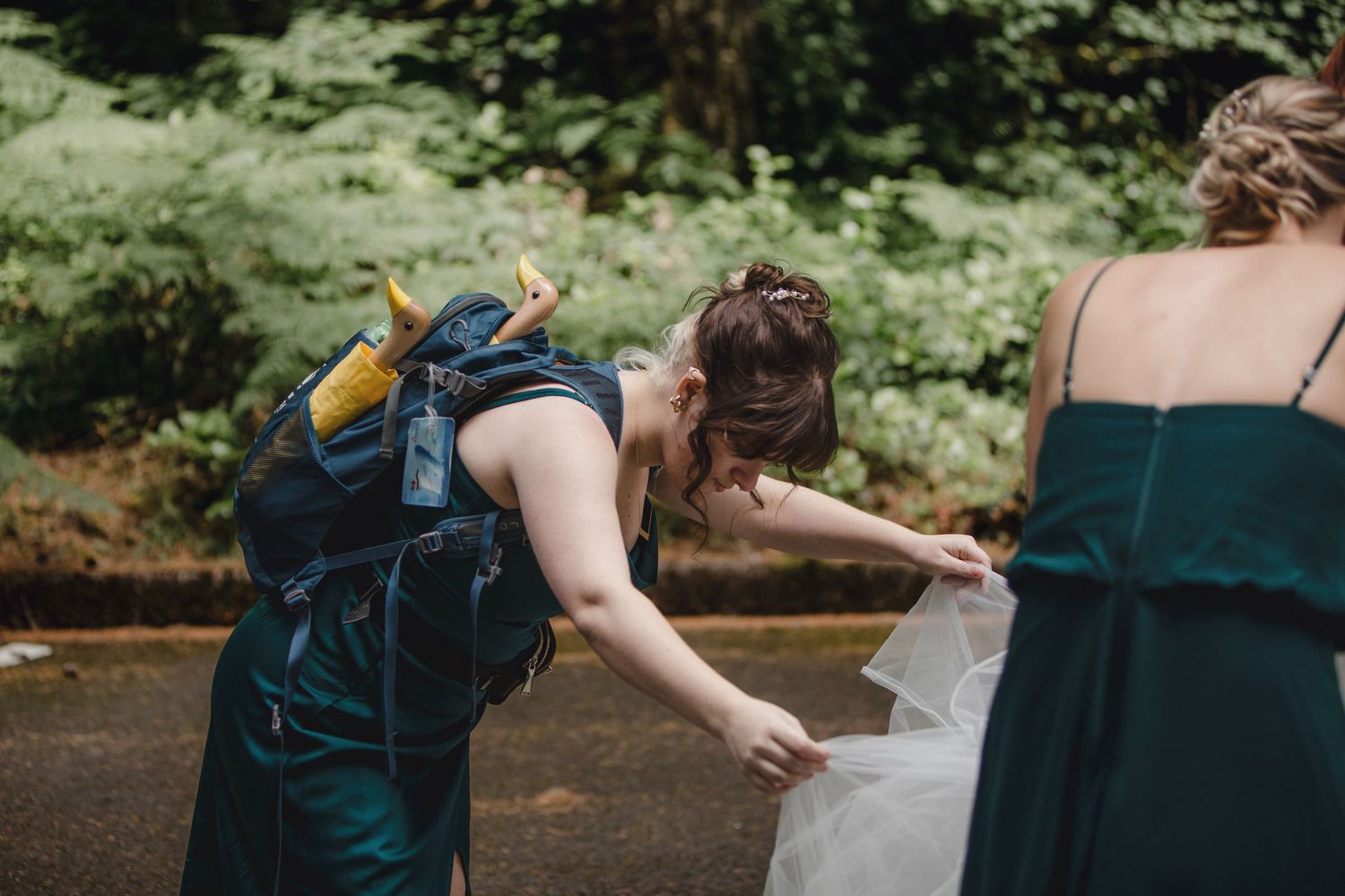 Someone helps adjust a long flowing dress outside in a woodsy setting while wearing a dark green formal gown.