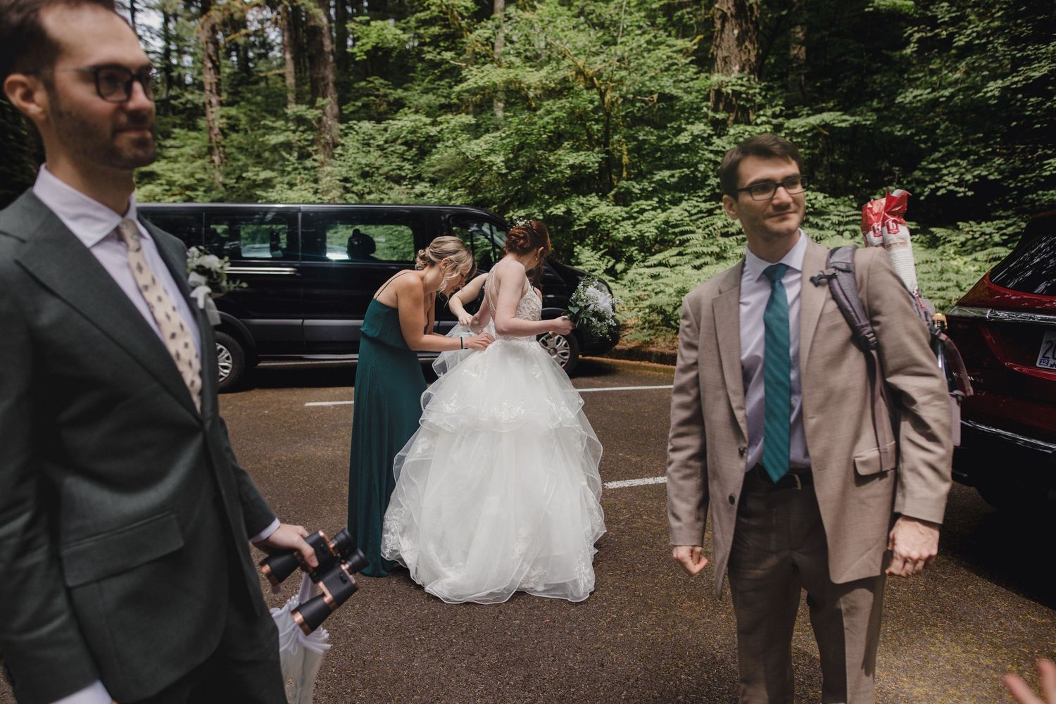 Wedding party members gather near parked cars on a forest path during an outdoor wedding celebration.
