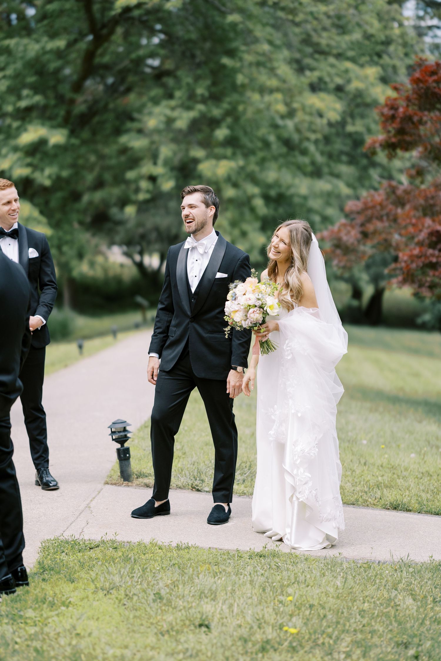 Wedding party members walking along a garden path surrounded by lush greenery on a summer day at Silver Bay YMCA in Lake George, NY.