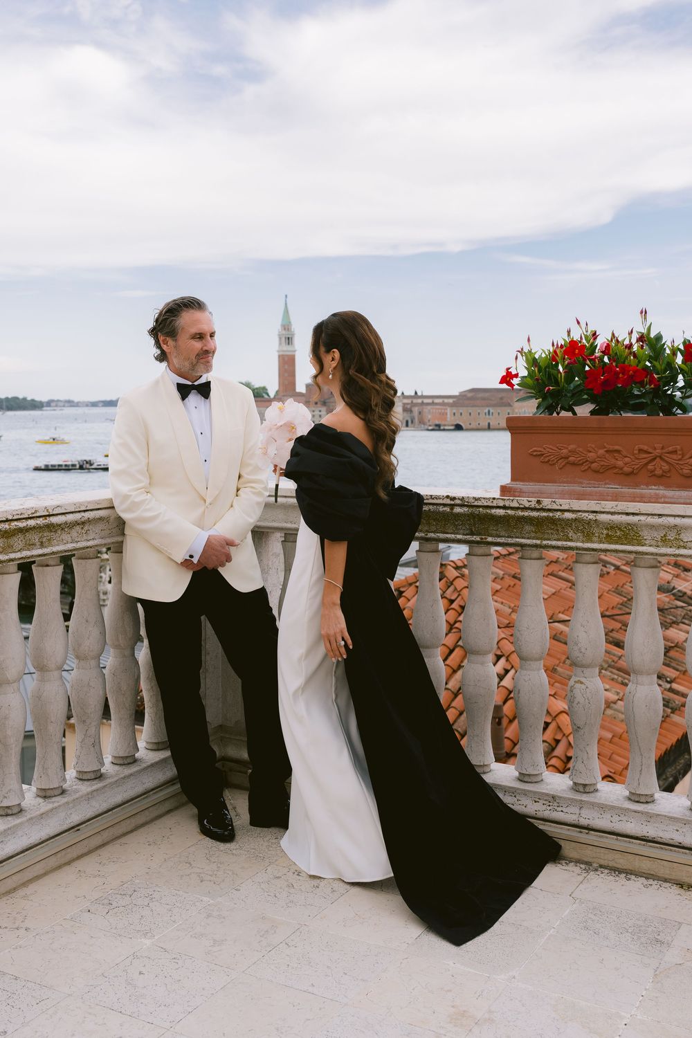 Elegant couple in formal attire share intimate moments on a balcony overlooking a scenic cityscape. Photoshoot by Venice Photographer Alina