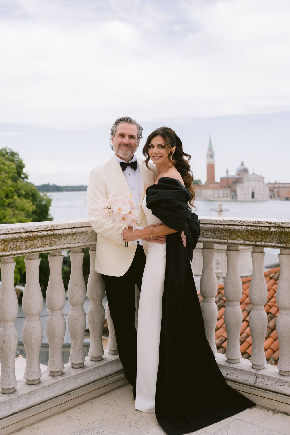 Romantic couple in black and white formal wear embrace on an ornate balcony with city views. Photoshoot by Venice Photographer Alina Indi