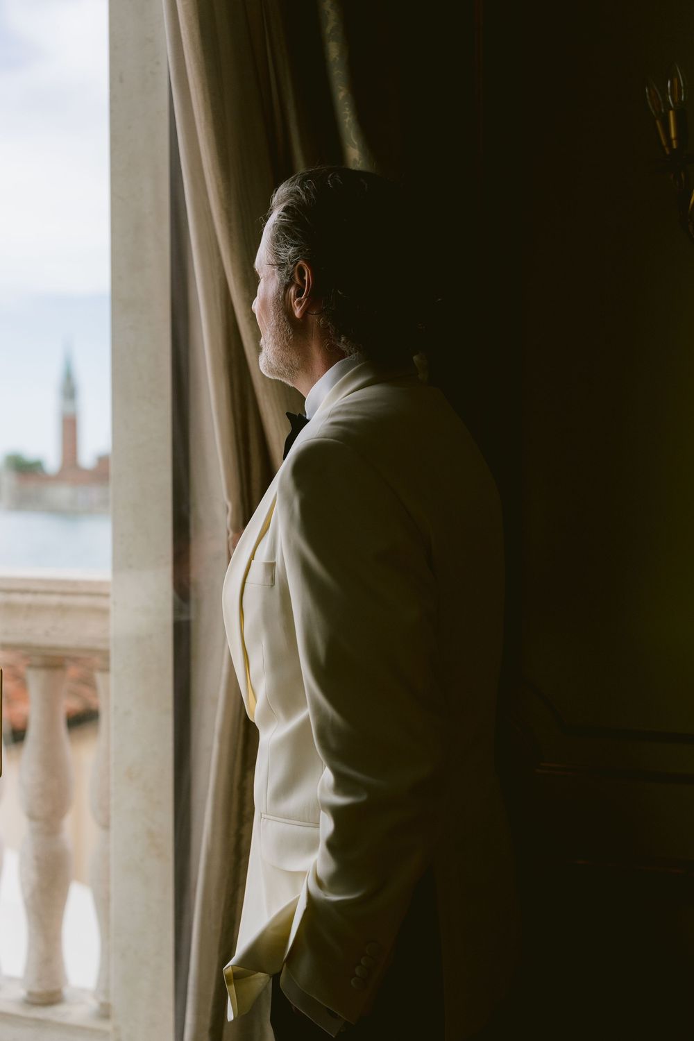 Silhouette of person in white jacket looking thoughtfully through window drapes. Photoshoot by Venice Photographer Alina Indi