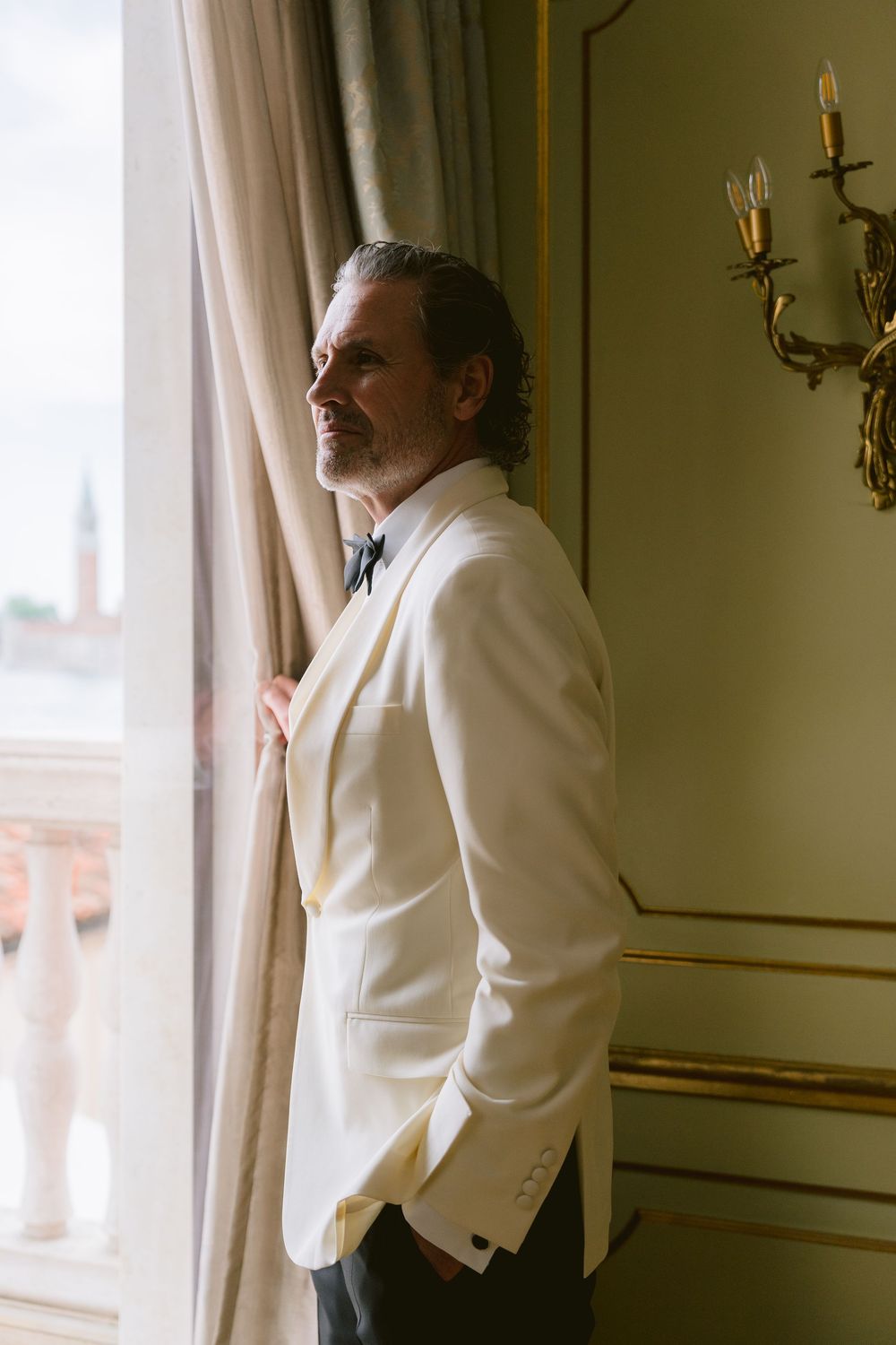 Person in white tuxedo jacket gazes out window in elegant interior setting. Photoshoot by Venice Photographer Alina Indi