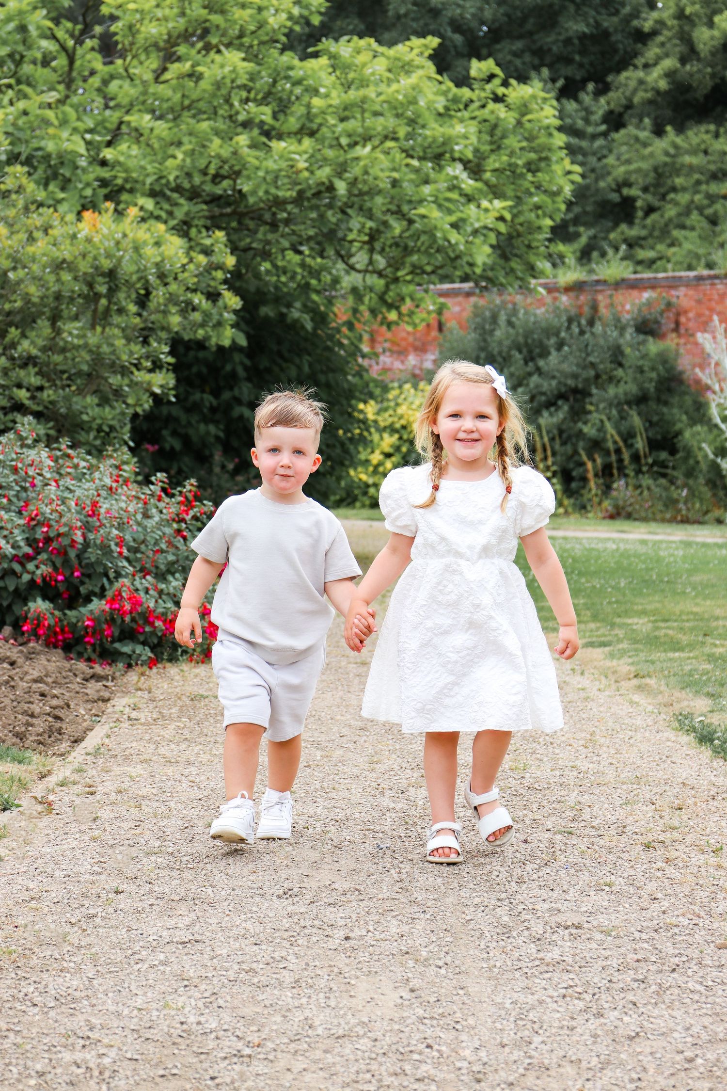 Two children in white outfits walk hand-in-hand down a garden path on a sunny summer day, during family shoot in Nottingham