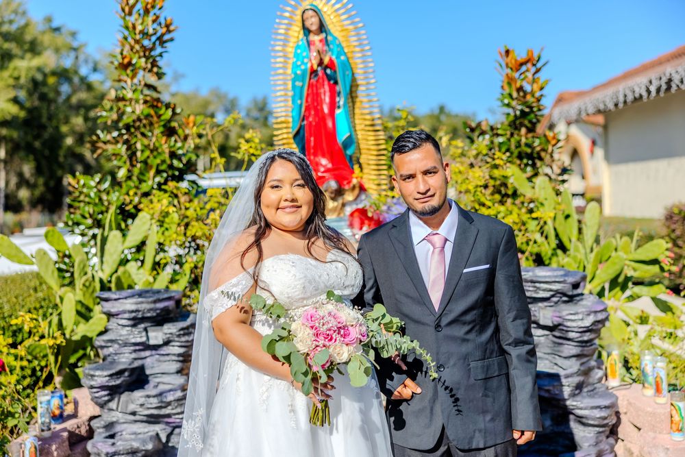 Wedding party poses outdoors with religious statues and pink floral decor in a garden setting.