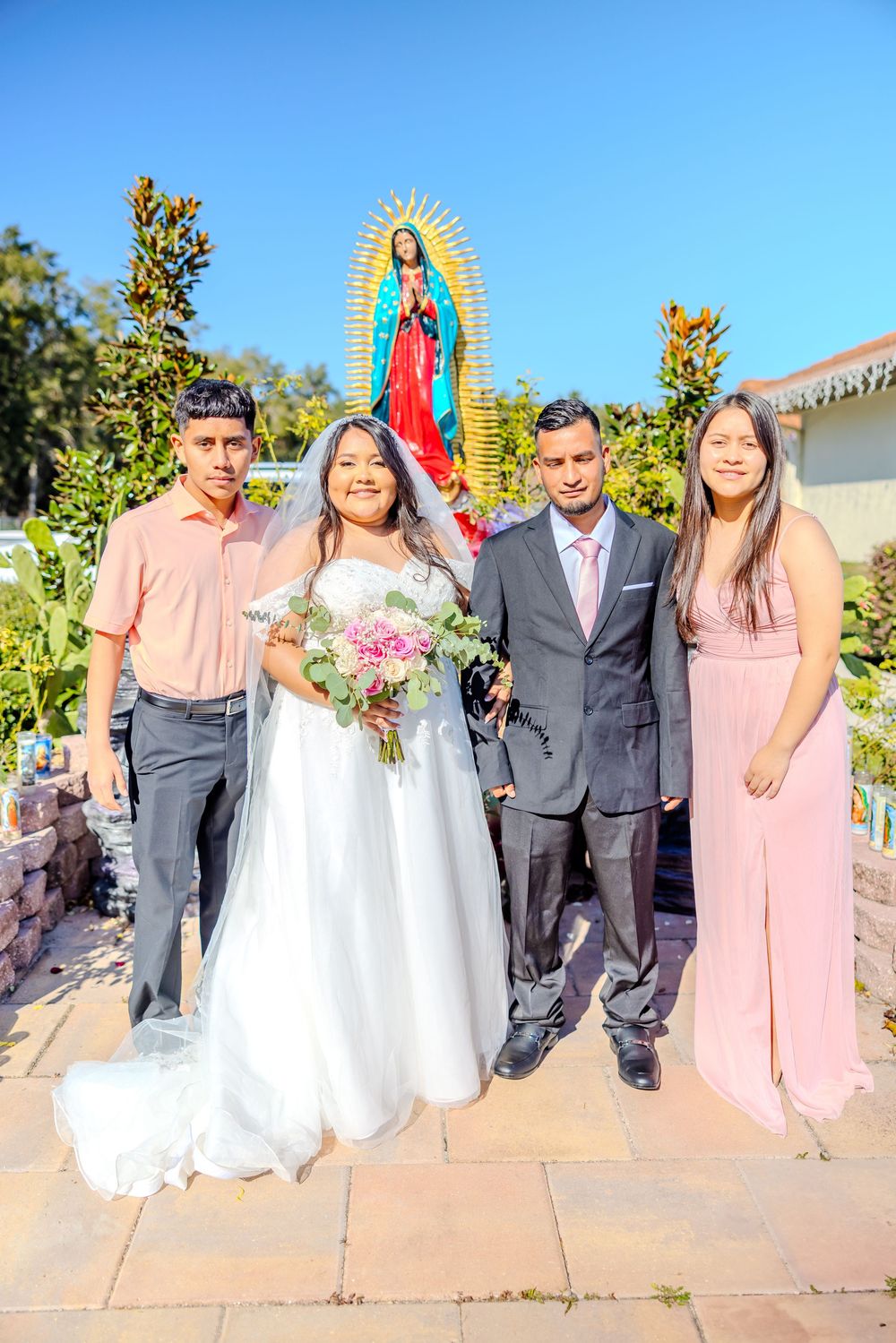 Wedding group portrait on stone path with religious icons and greenery in background.