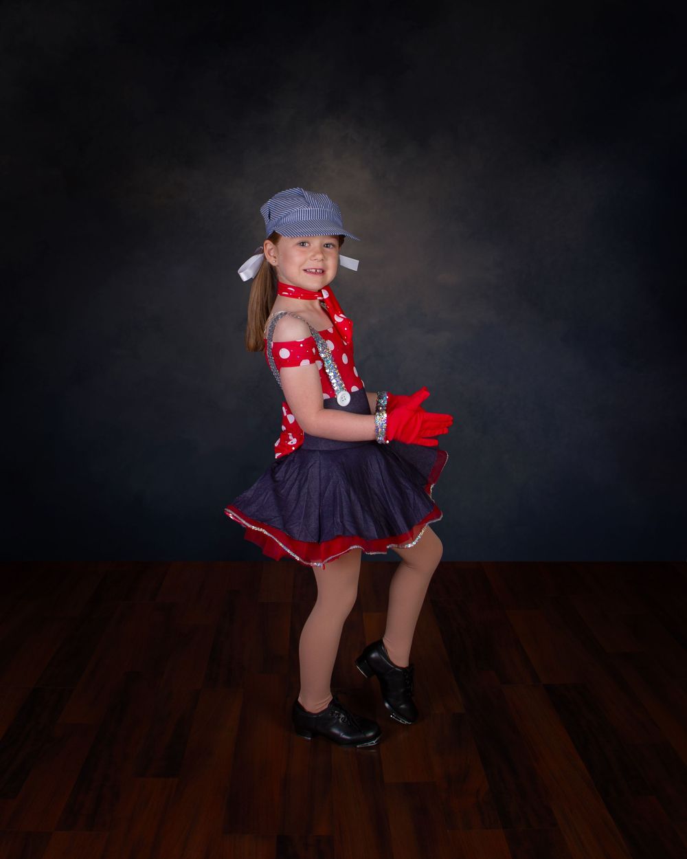 Young performer in red polka dot top, navy skirt and baseball cap poses during dance recital on dark stage.
