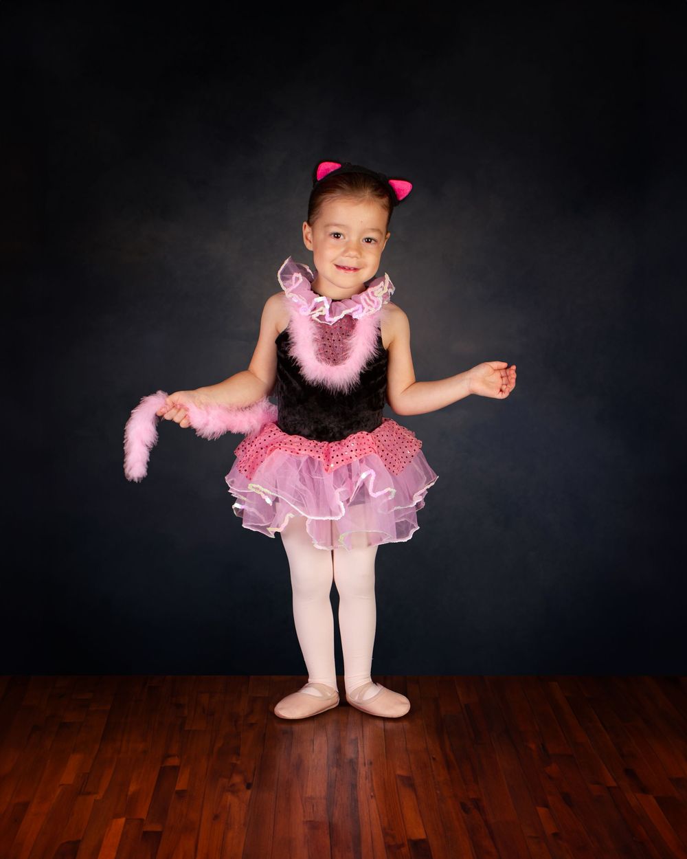 Young dancer in a pink and black tutu costume with matching bow poses on hardwood floor against dark backdrop.