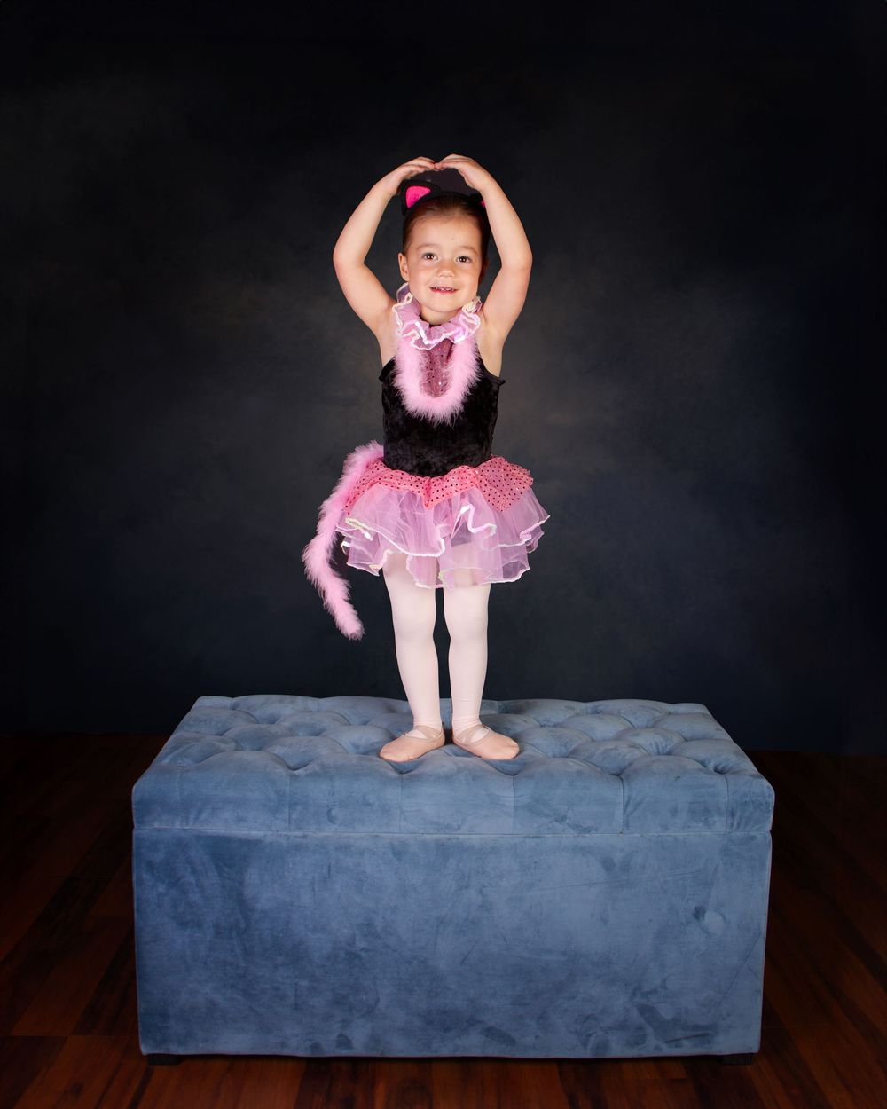 Young ballerina in pink tutu and white tights poses with arms raised while standing on blue cushioned platform.