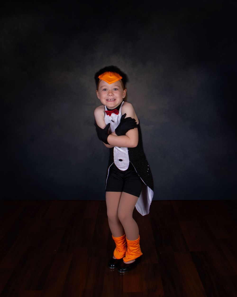Young performer in black dance costume with orange shoes strikes a playful pose against dark background on wooden floor.