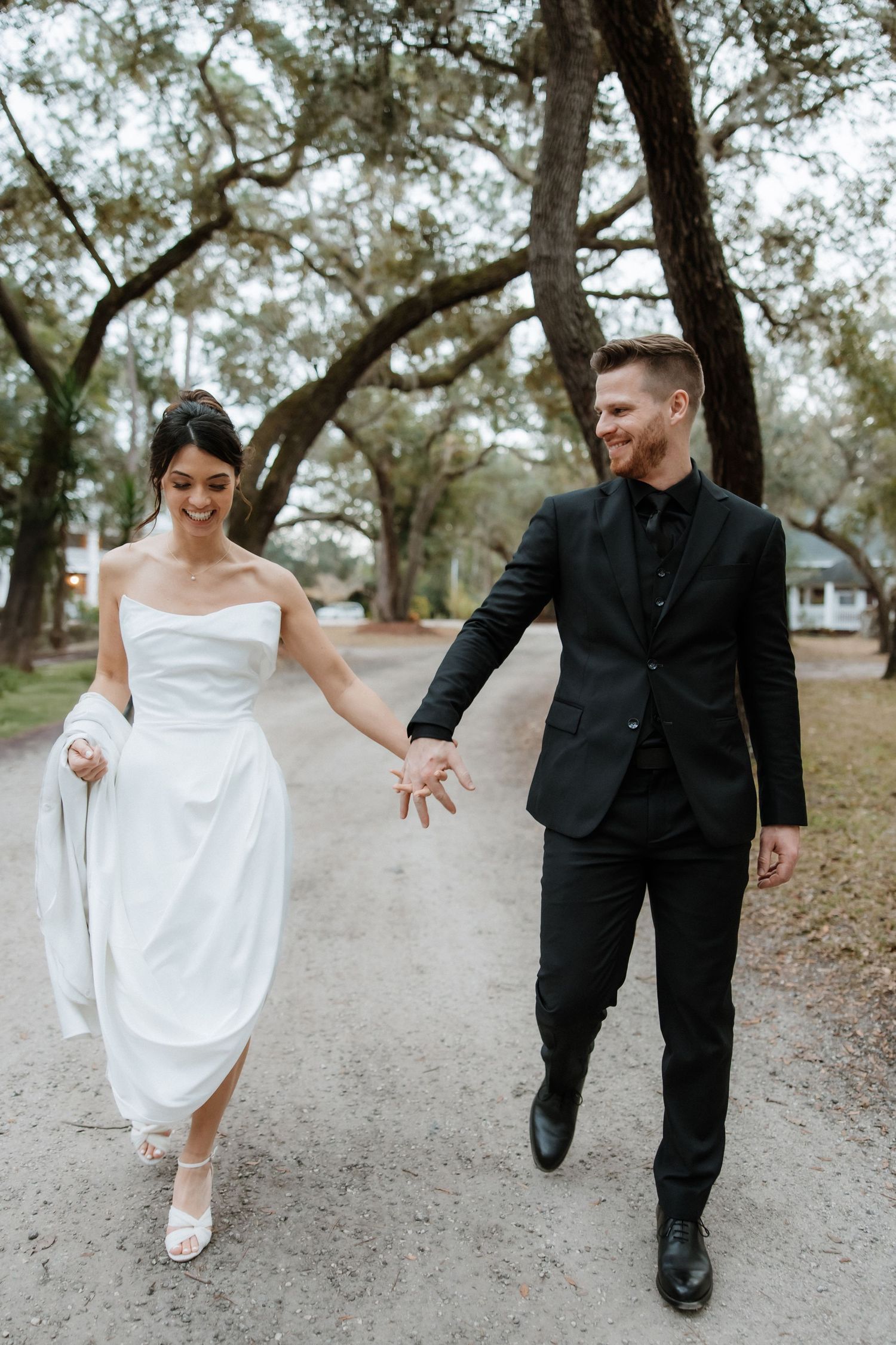 A couple in wedding attire walk hand in hand down a tree-lined dirt path in a romantic outdoor setting.