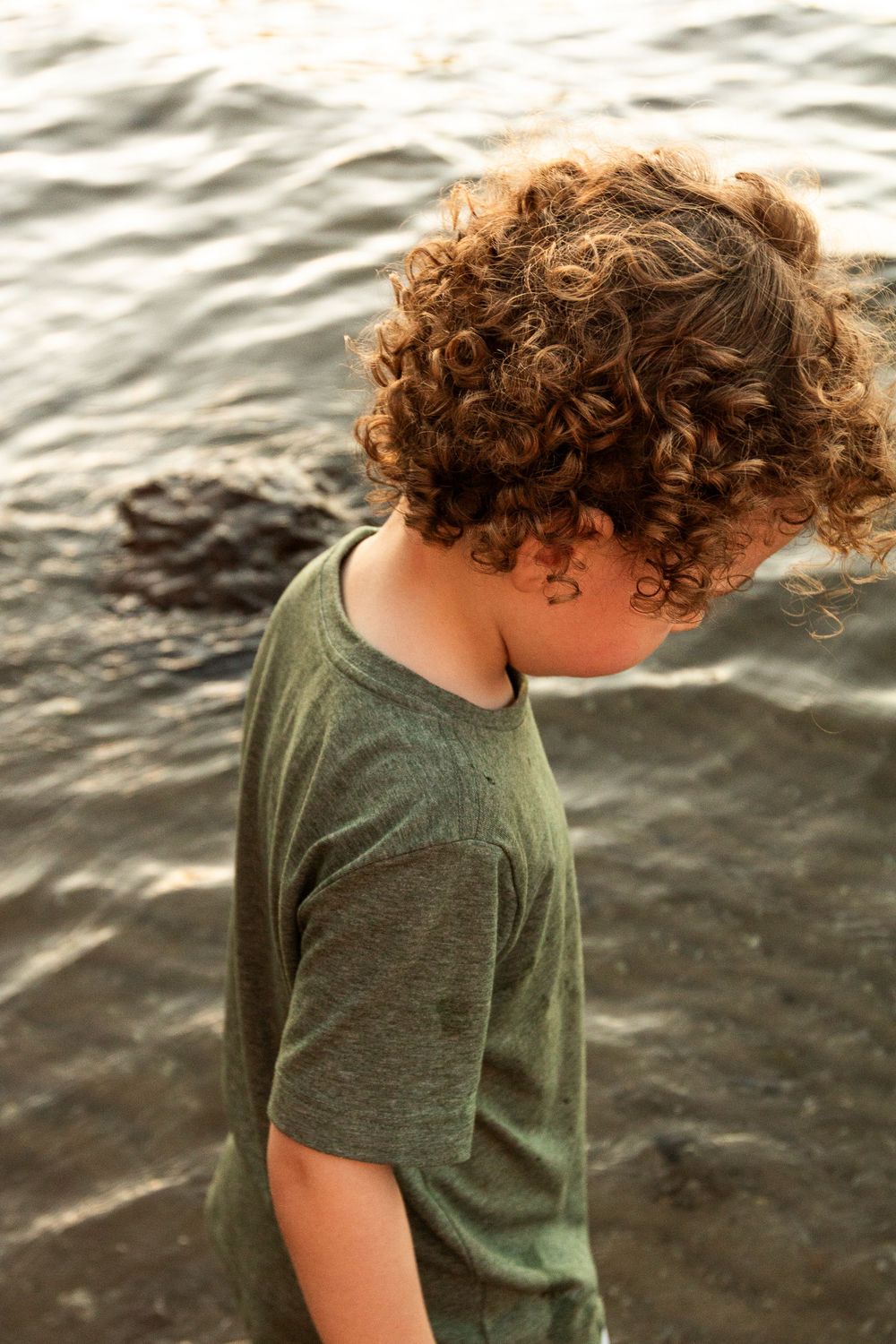 Profile view of curly-haired figure in olive green t-shirt against rippling water background.