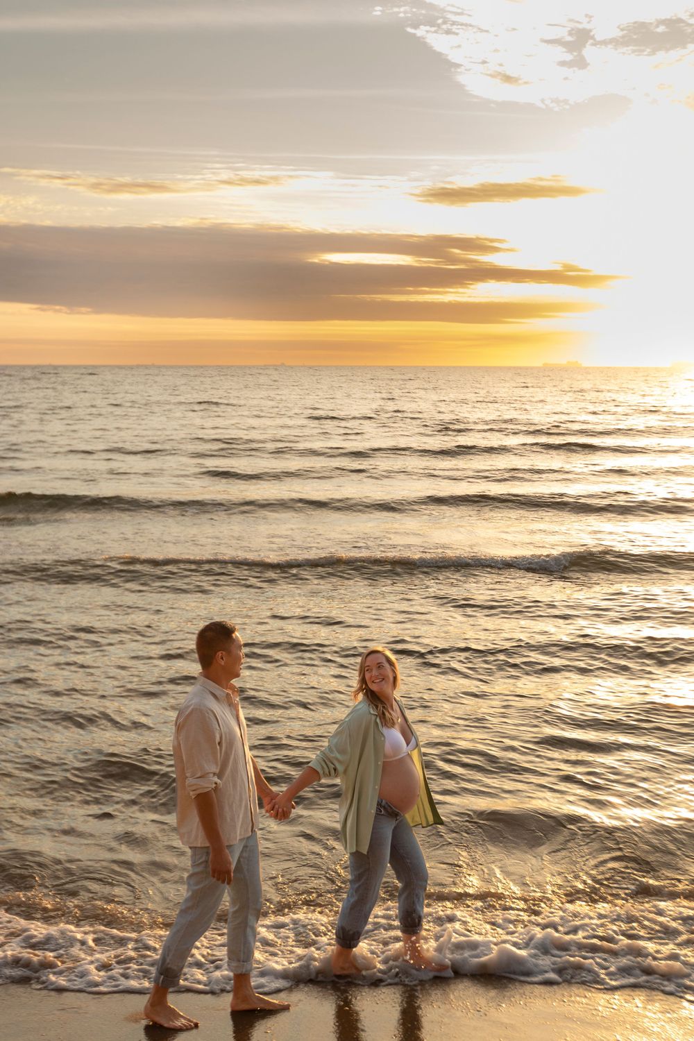 Couple walking along beach shoreline at sunset as waves crash against their feet.