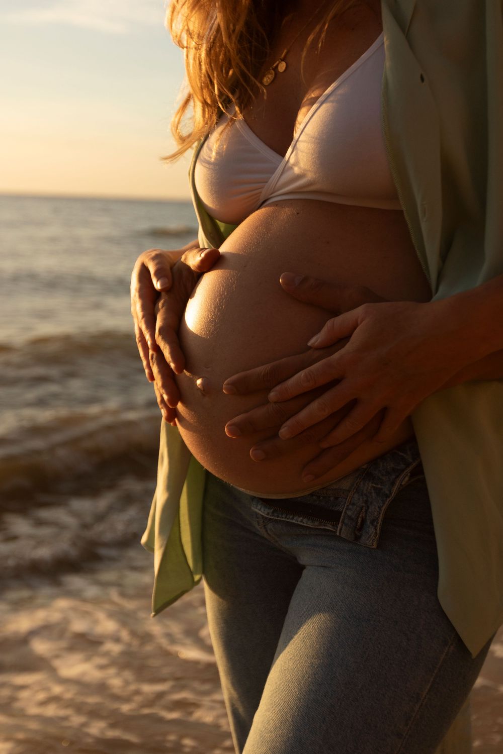 Close-up of pregnant belly with hands resting on it during sunset beach photoshoot.