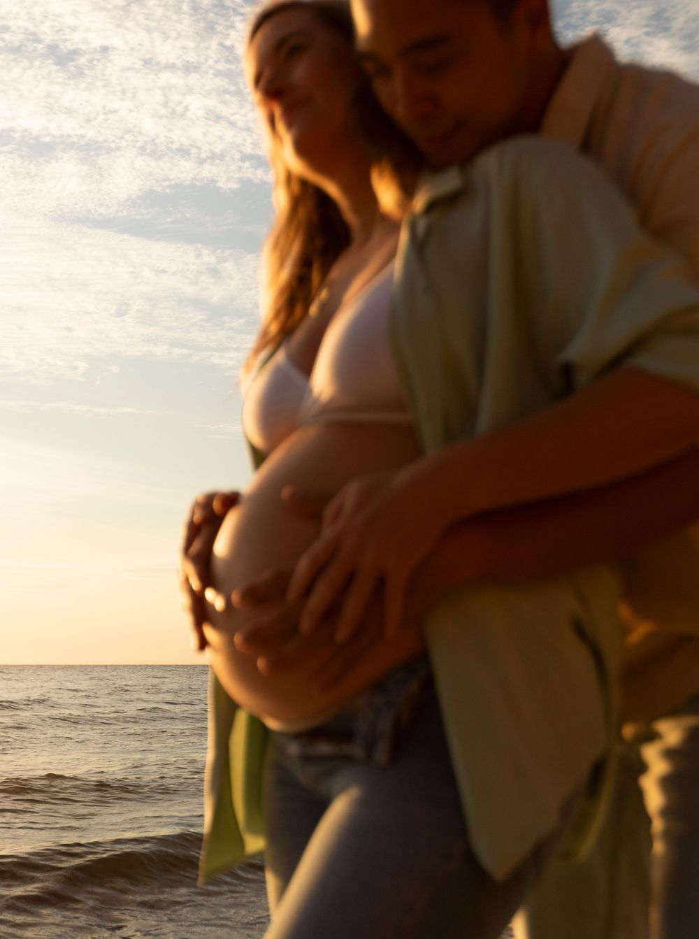 Intimate beach sunset photo of couple embracing with focus on pregnant belly.