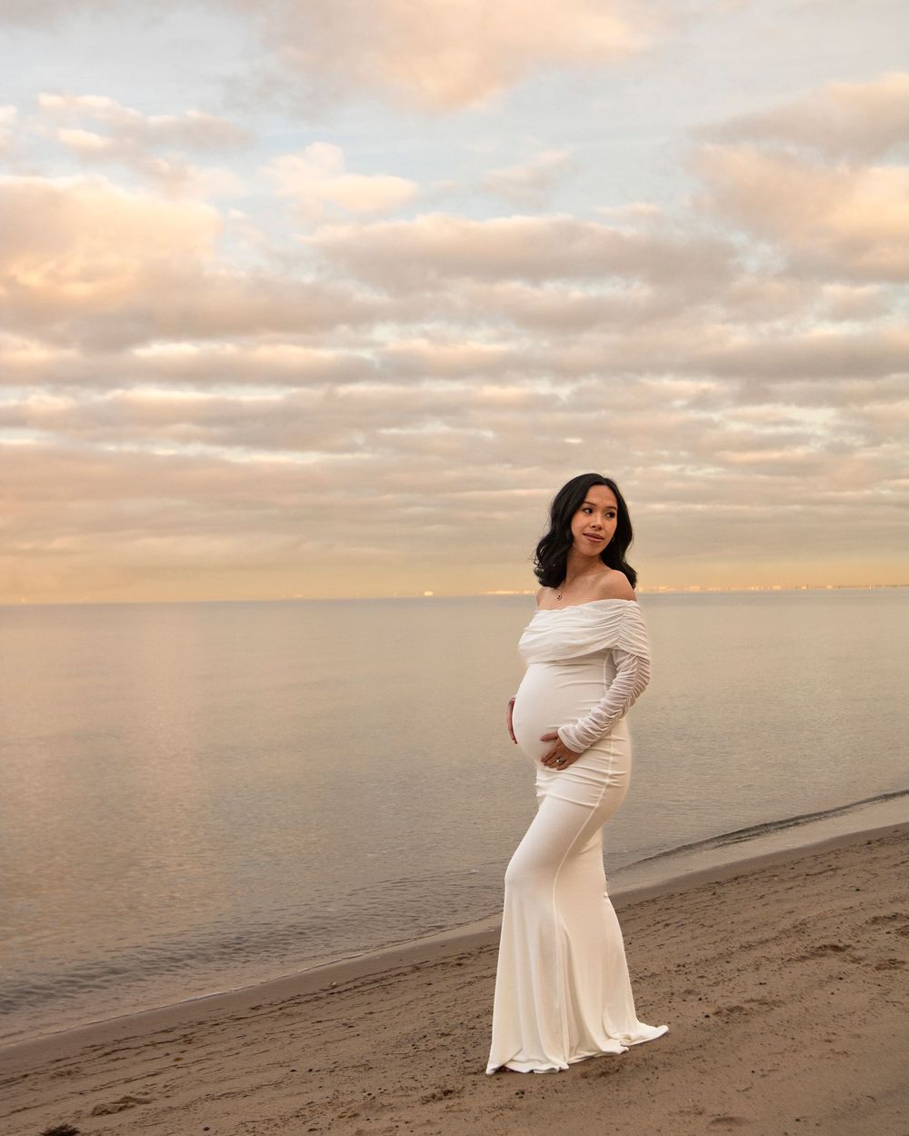 A maternity photo of a woman in a white dress standing on a beach at sunset.