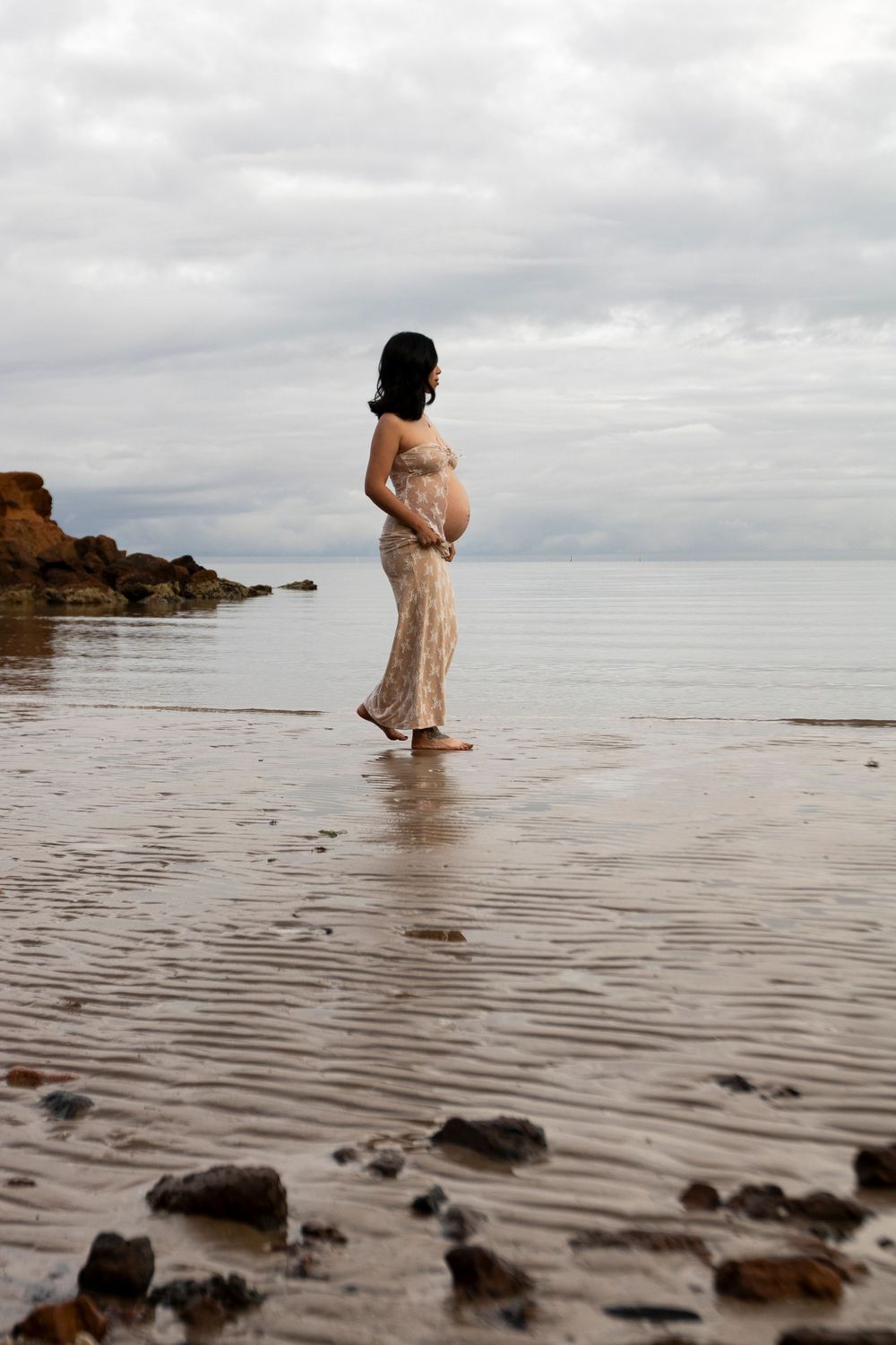 An expectant mother standing on wet sand by rocky coastline for a maternity shoot.