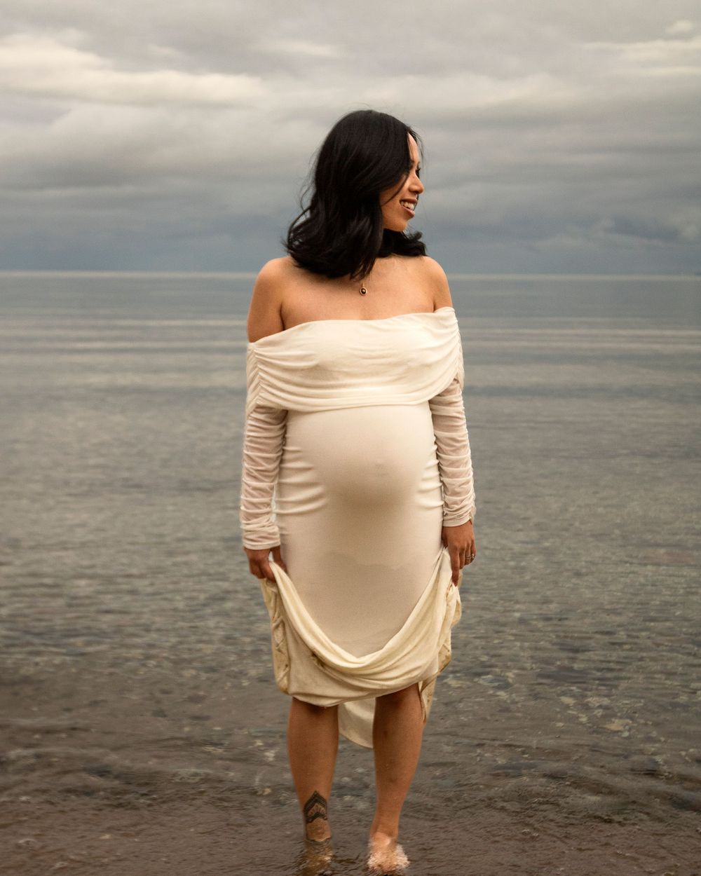 Woman in off-shoulder cream dress standing at water's edge during cloudy beach photoshoot.