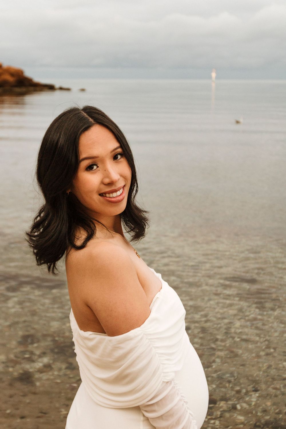 Candid beach maternity photo with woman in white off-shoulder dress near rocky shore.