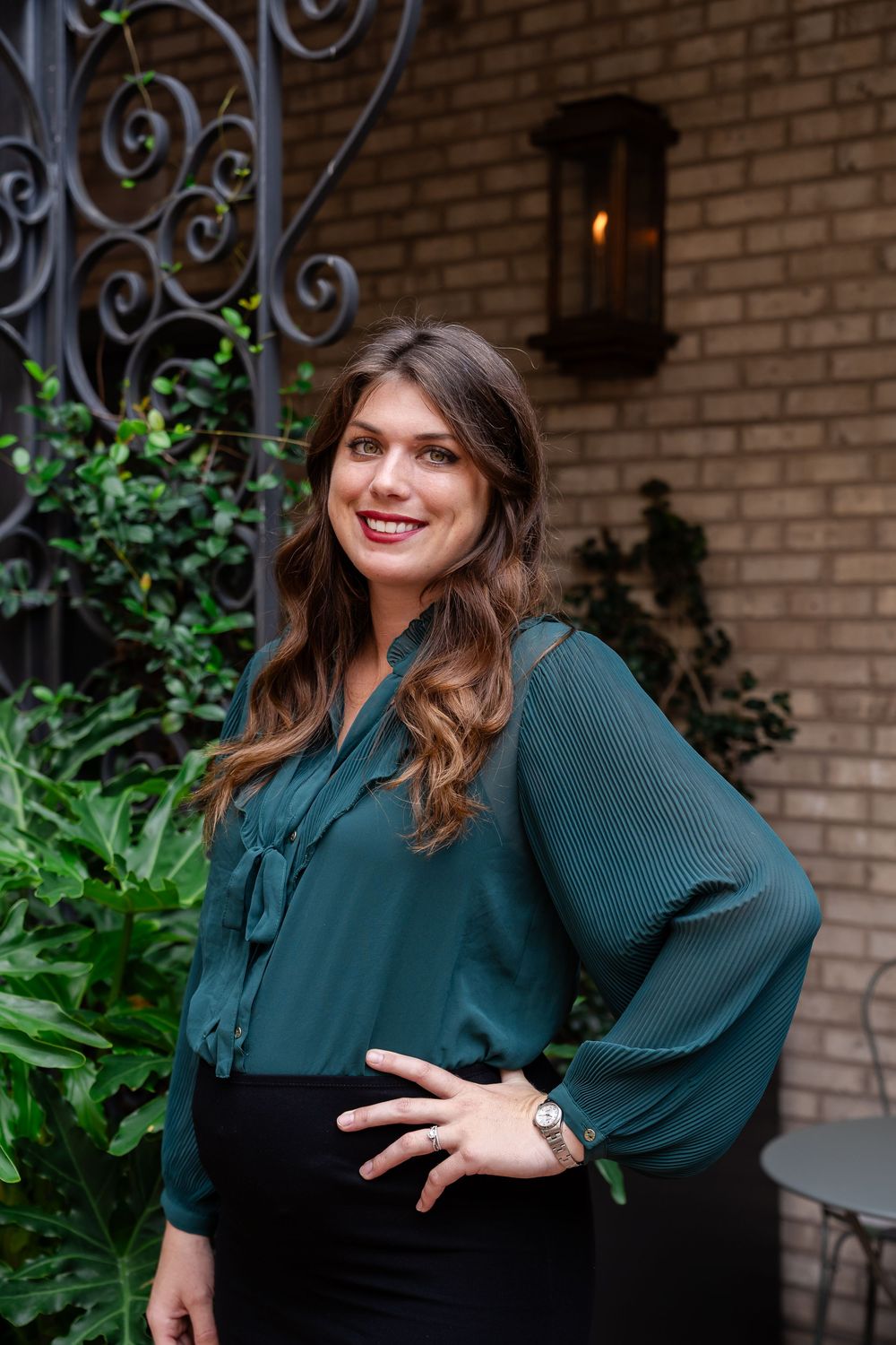 Professional portrait of woman in teal blouse against brick wall with decorative ironwork.