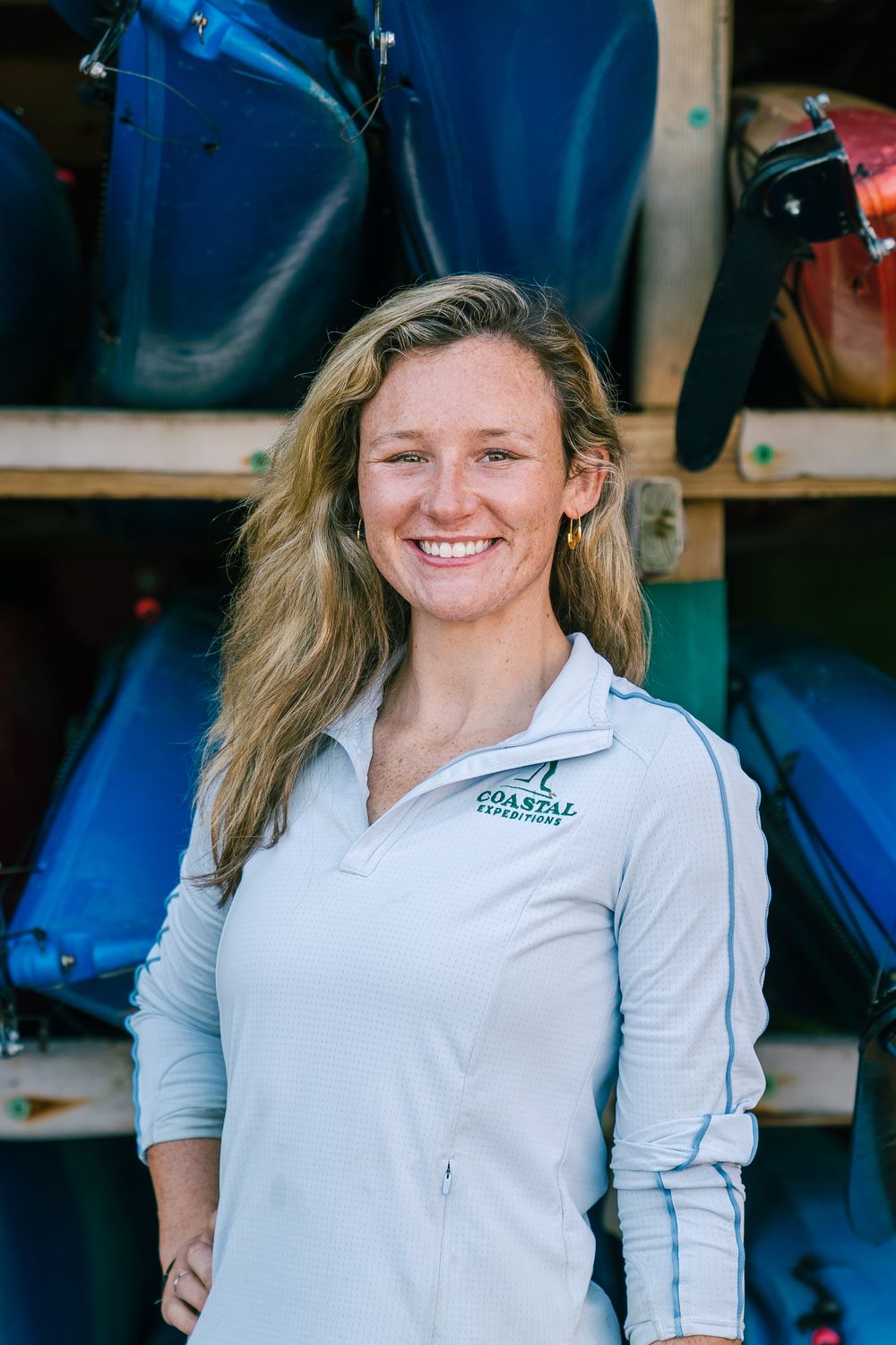 Staff member poses in front of rack of blue kayaks at outdoor recreation facility.