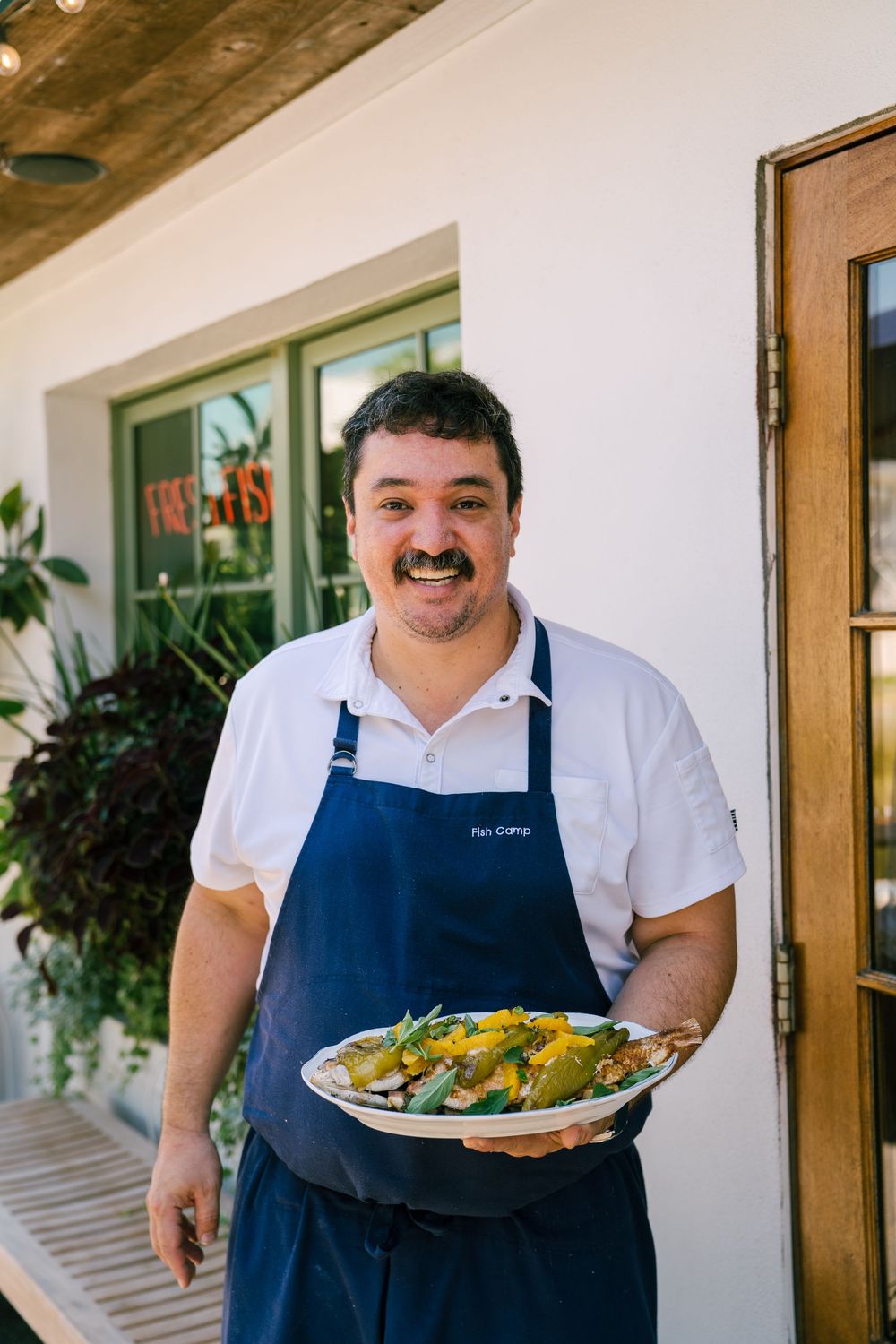 Staff member in blue apron presents plated meal outside restaurant entrance.