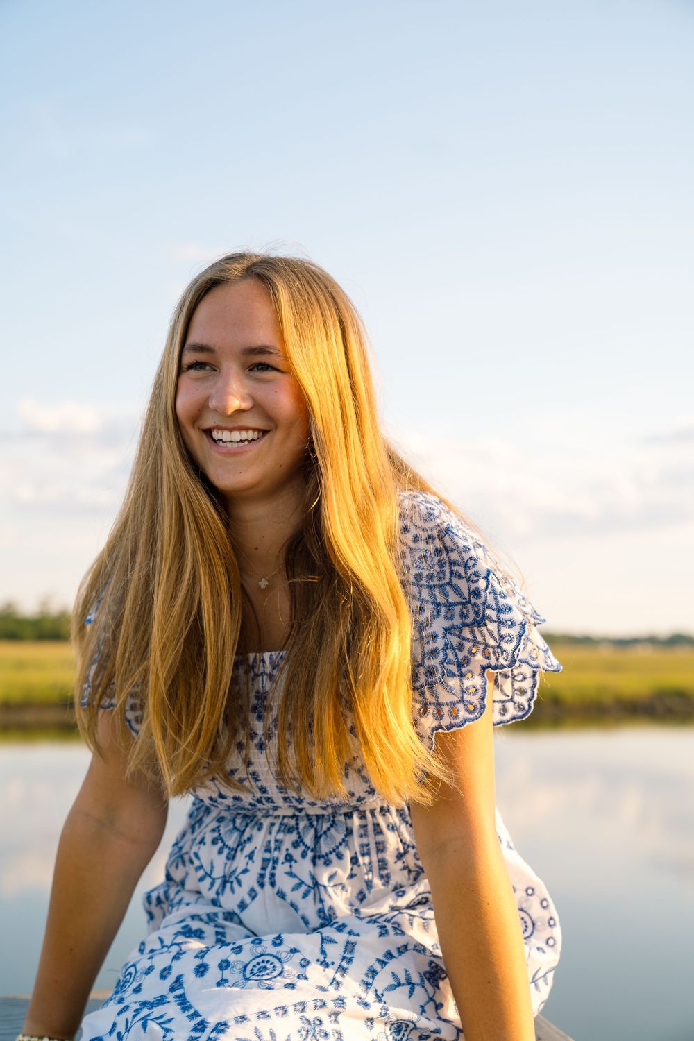 Woman in blue and white patterned dress smiling outdoors at sunset near water.