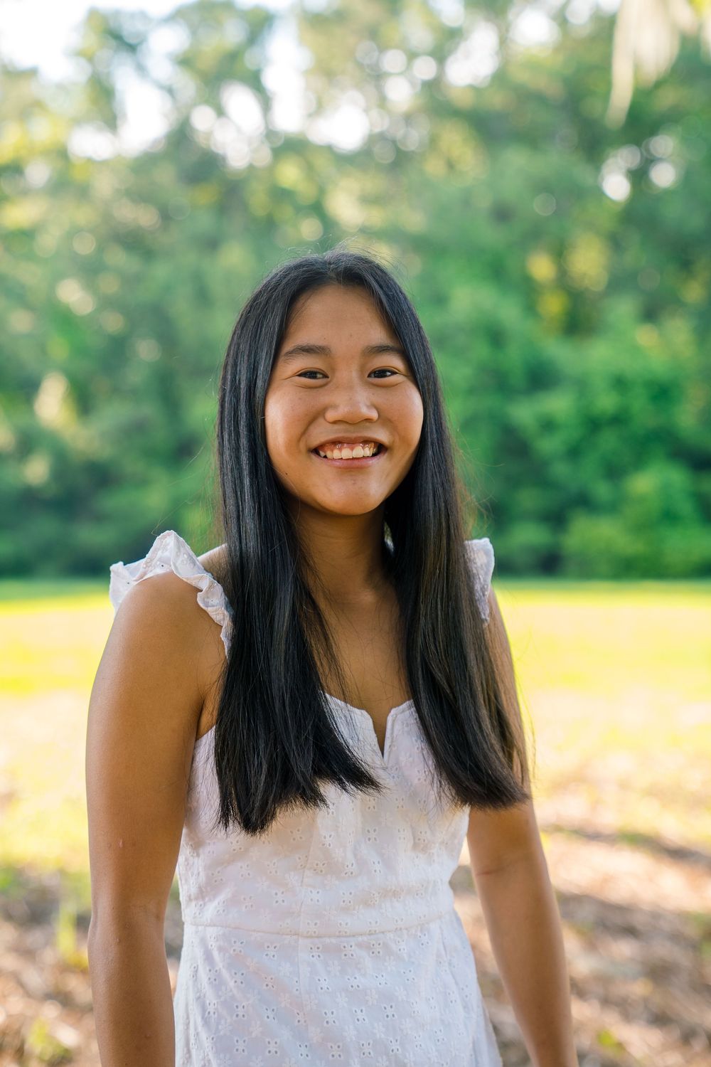Woman in white eyelet dress smiling outdoors in a sunny natural setting.