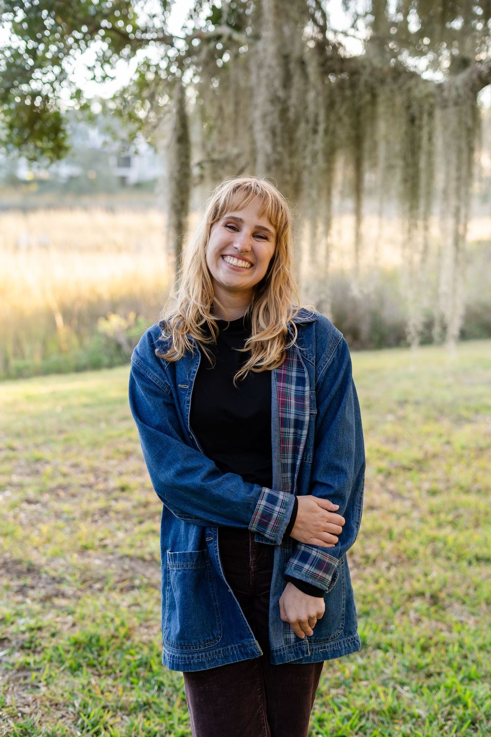 Person wearing denim jacket poses outdoors in field with Spanish moss trees in background at golden hour.