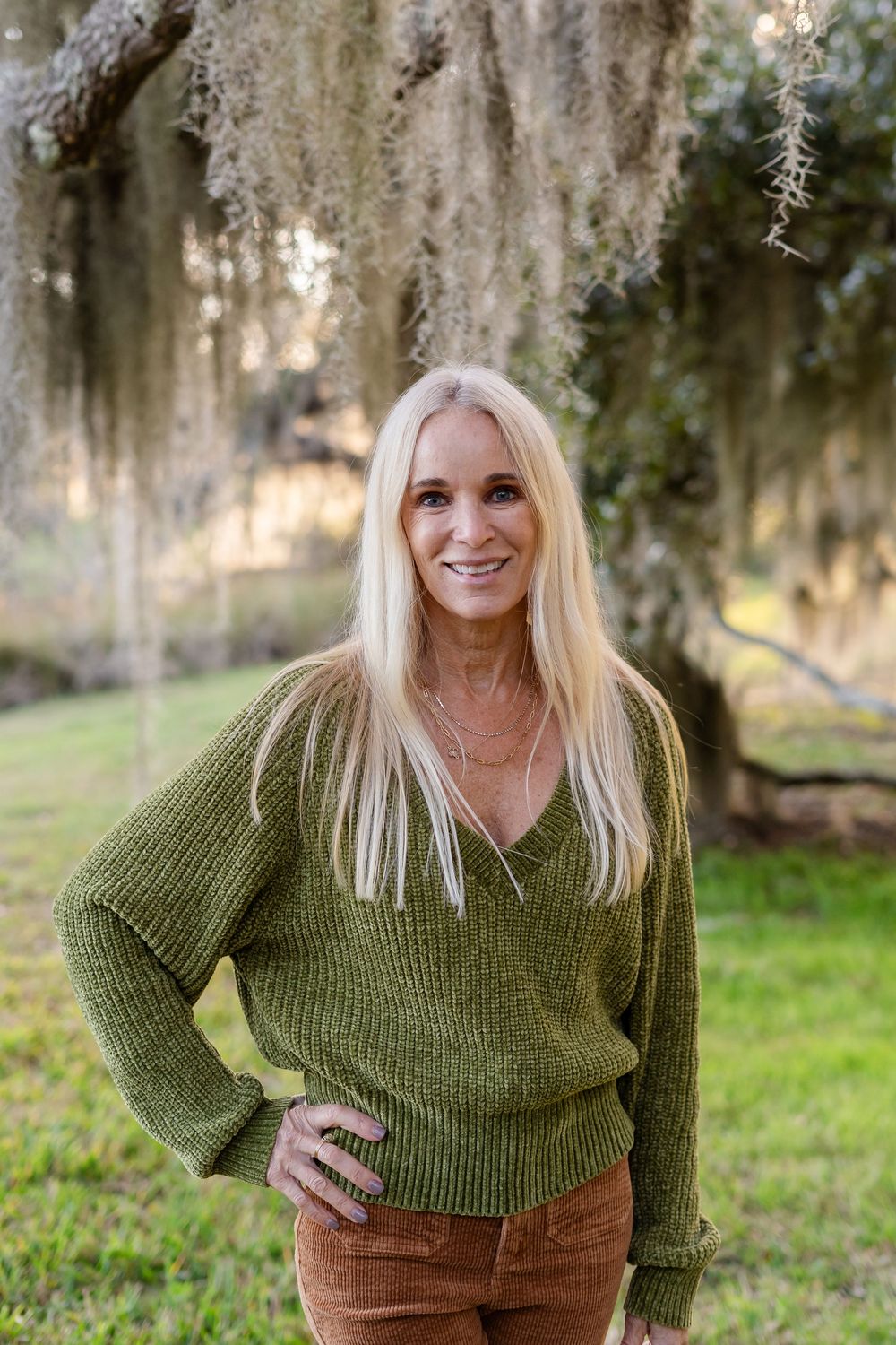 A person in a green knit sweater poses outdoors against a backdrop of Spanish moss and trees.
