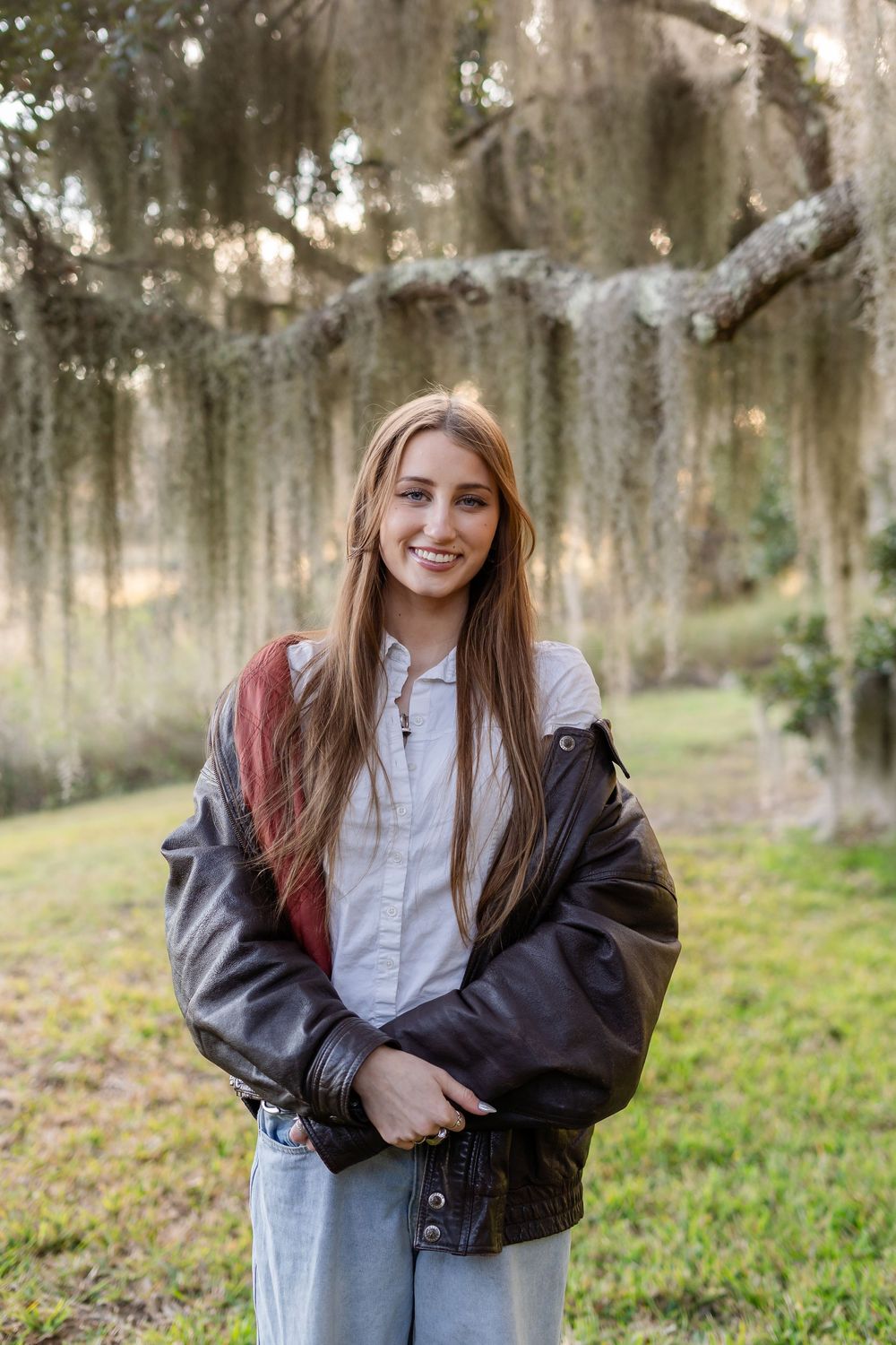 A person wearing a black jacket and white shirt poses outdoors with Spanish moss hanging from trees in the background.