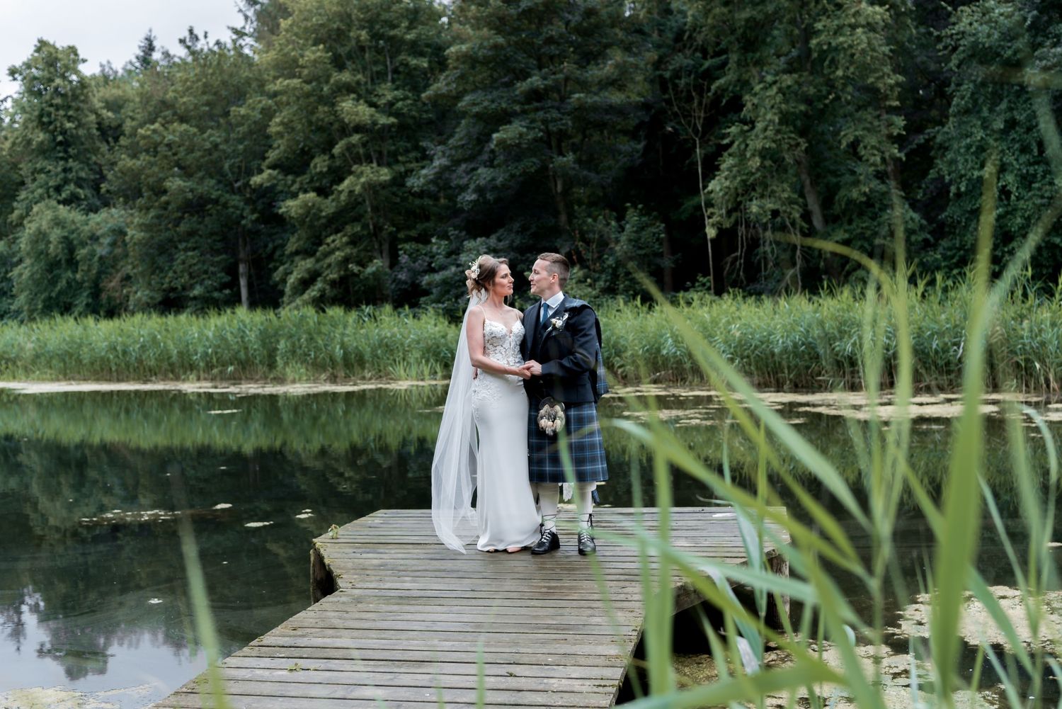 A couple in wedding attire stands together on a wooden dock overlooking a tranquil pond surrounded by lush greenery.