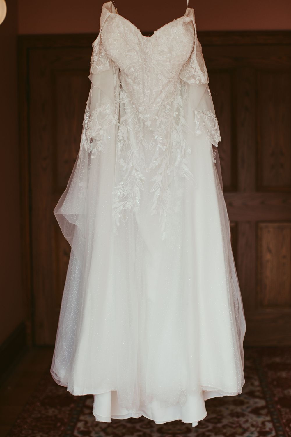 White wedding dress with lace details hanging on display against wooden backdrop.