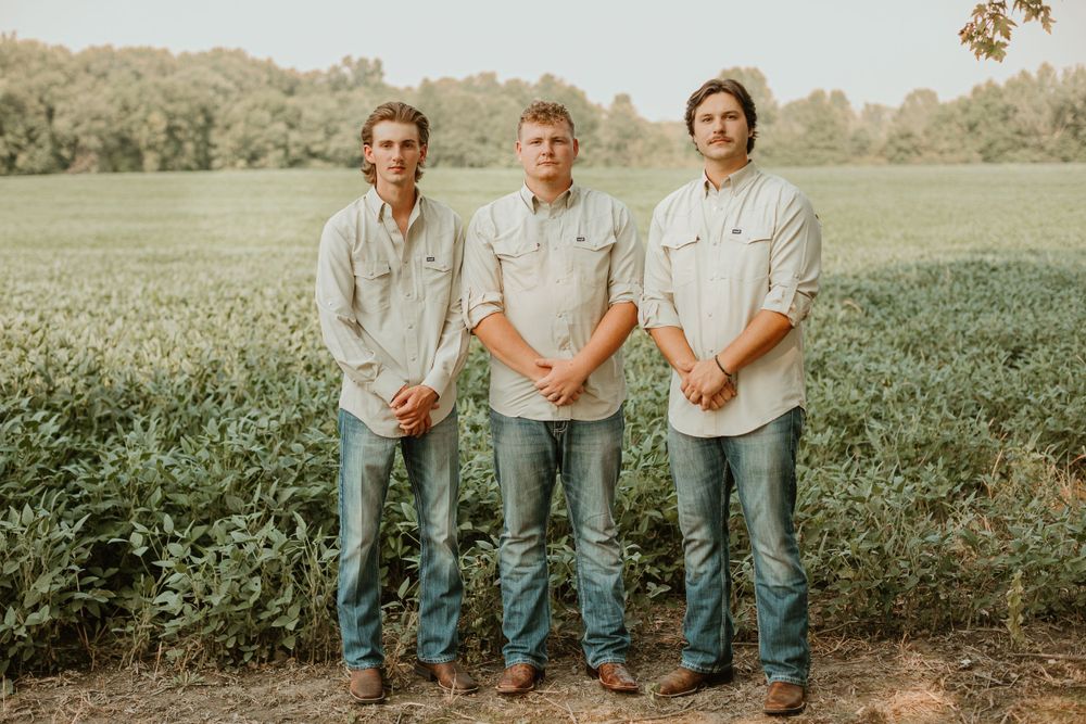 Three groomsmen in light shirts and jeans pose casually in a rustic outdoor setting.