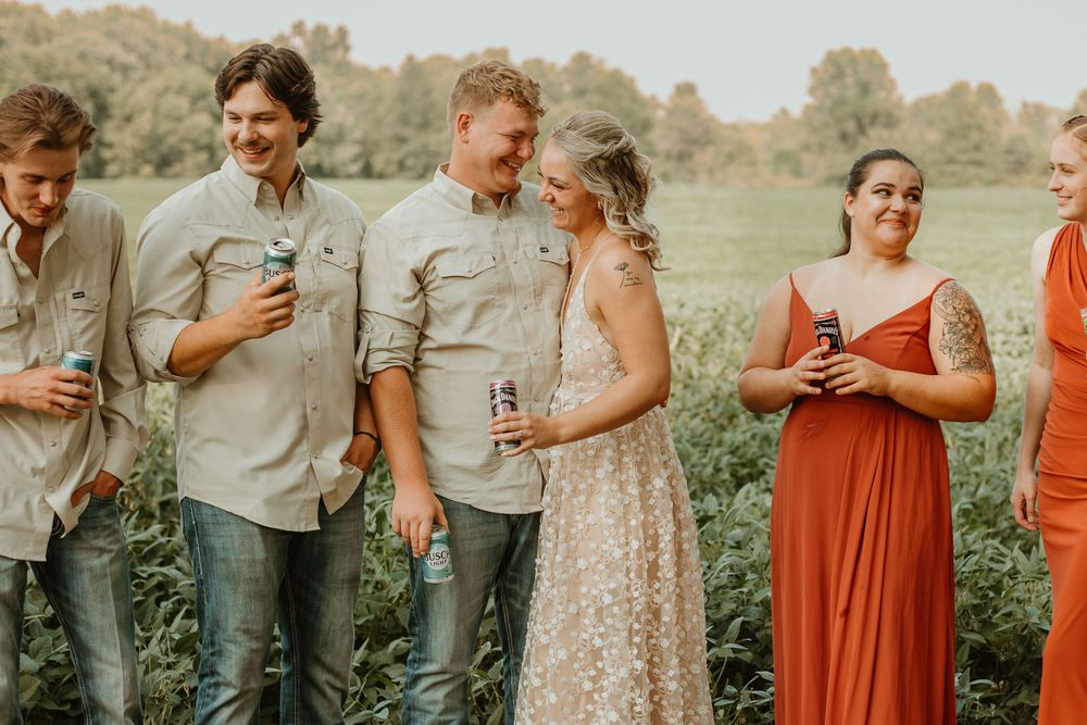 Wedding party members in coral dresses and casual attire pose together in a field.