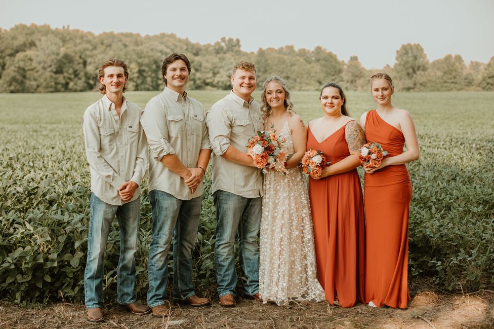 Wedding party lined up outdoors wearing coral dresses and light colored shirts.