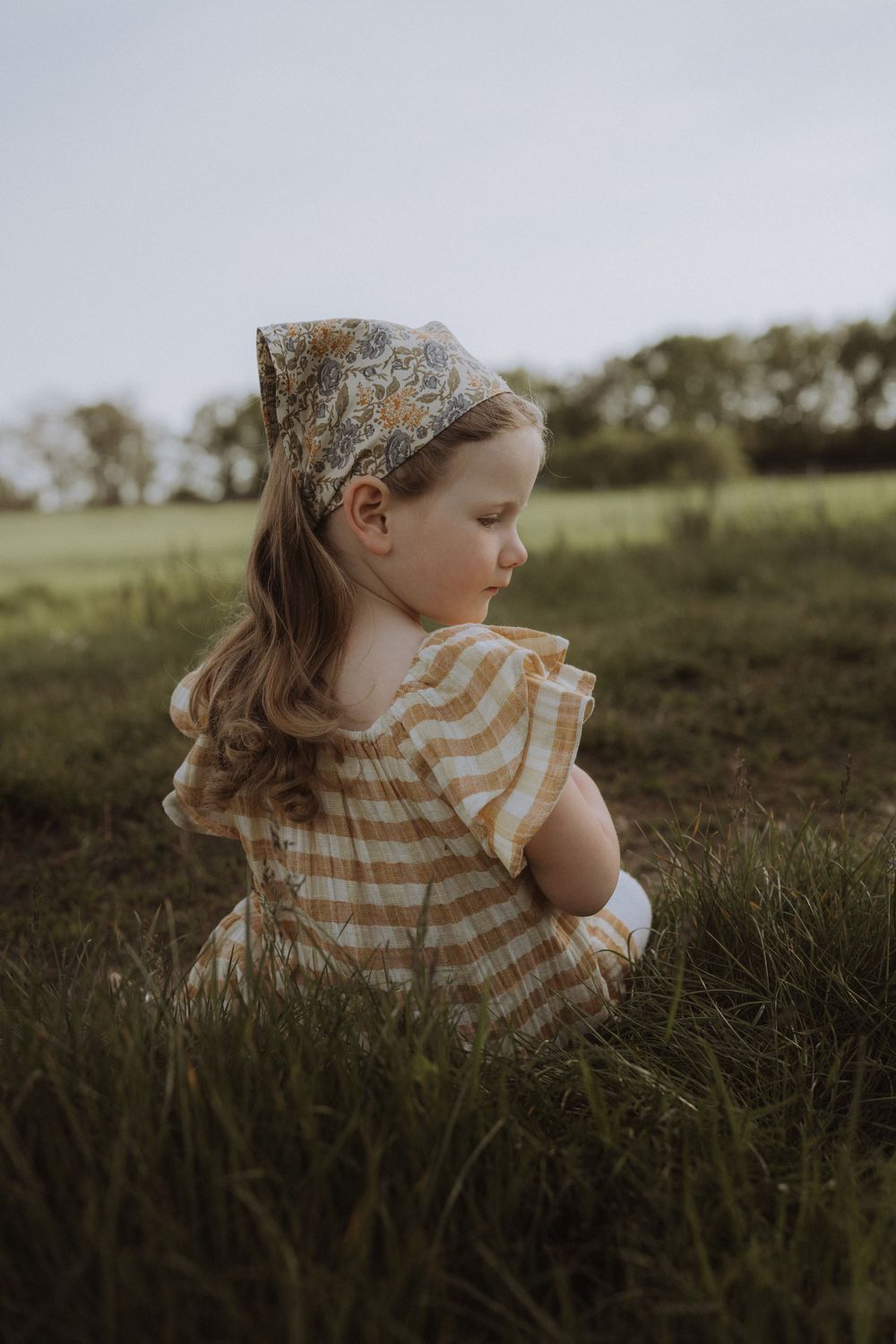 Young figure in striped dress and lace headband sits thoughtfully in a sunny meadow.