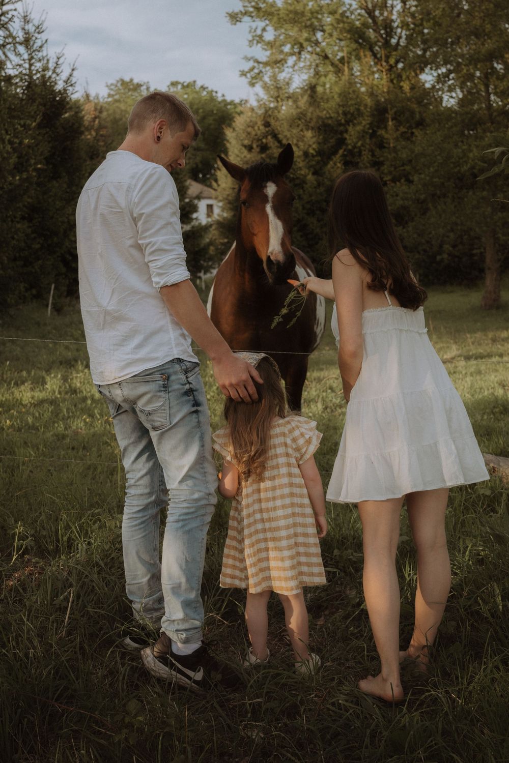 Family and horse standing together in field at sunset with child in gingham dress.