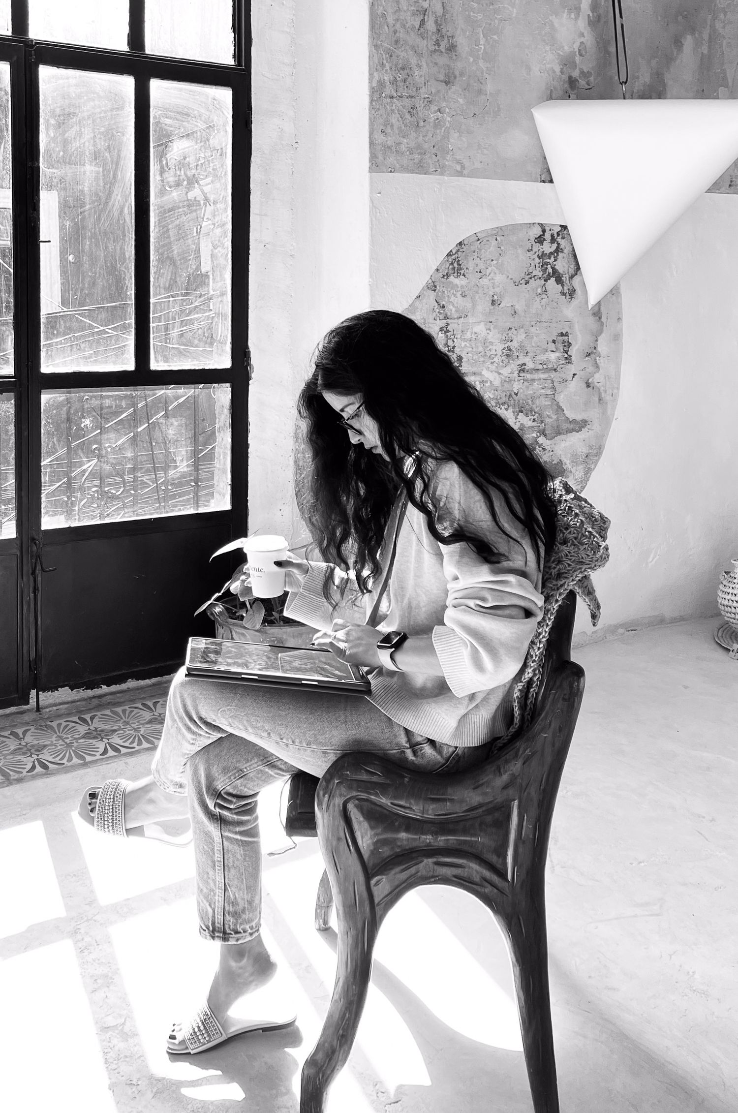 A person with long dark hair sits in a wooden chair reading a book by a window in black and white photography.