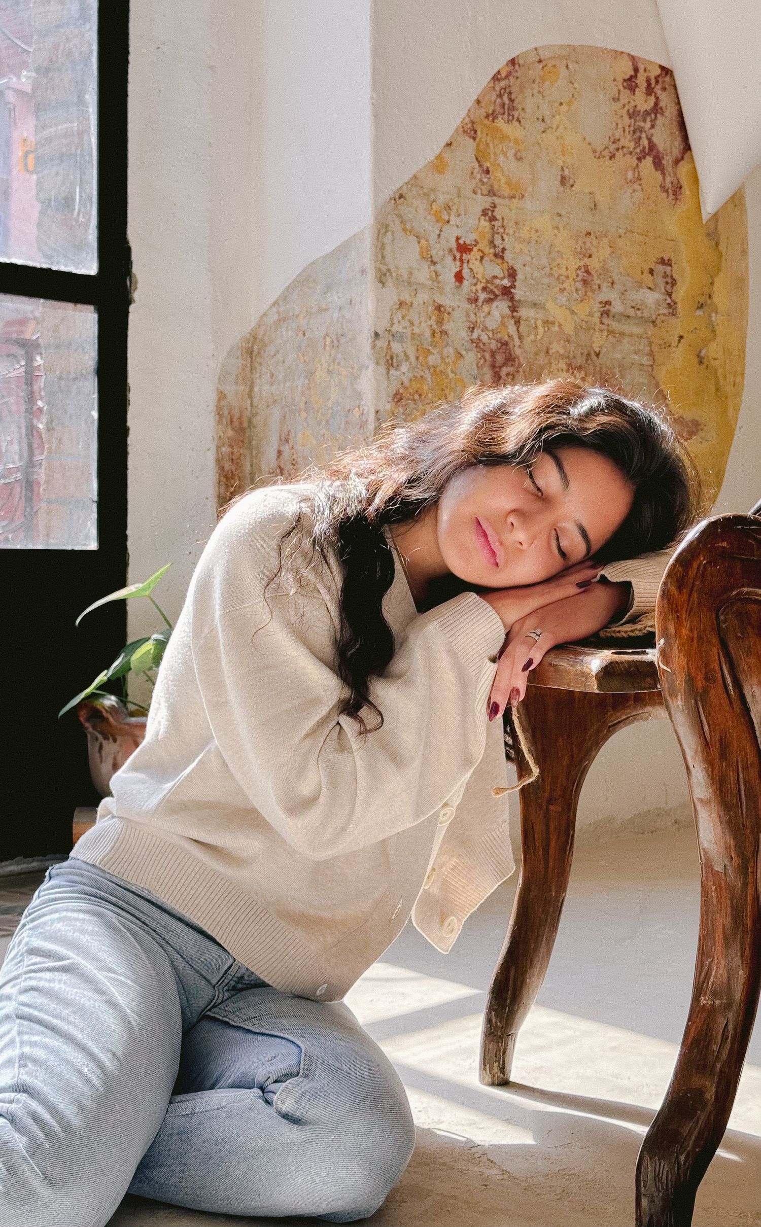 The person rests against a wooden chair near a window in a beige sweater and jeans, with sunlight streaming in.