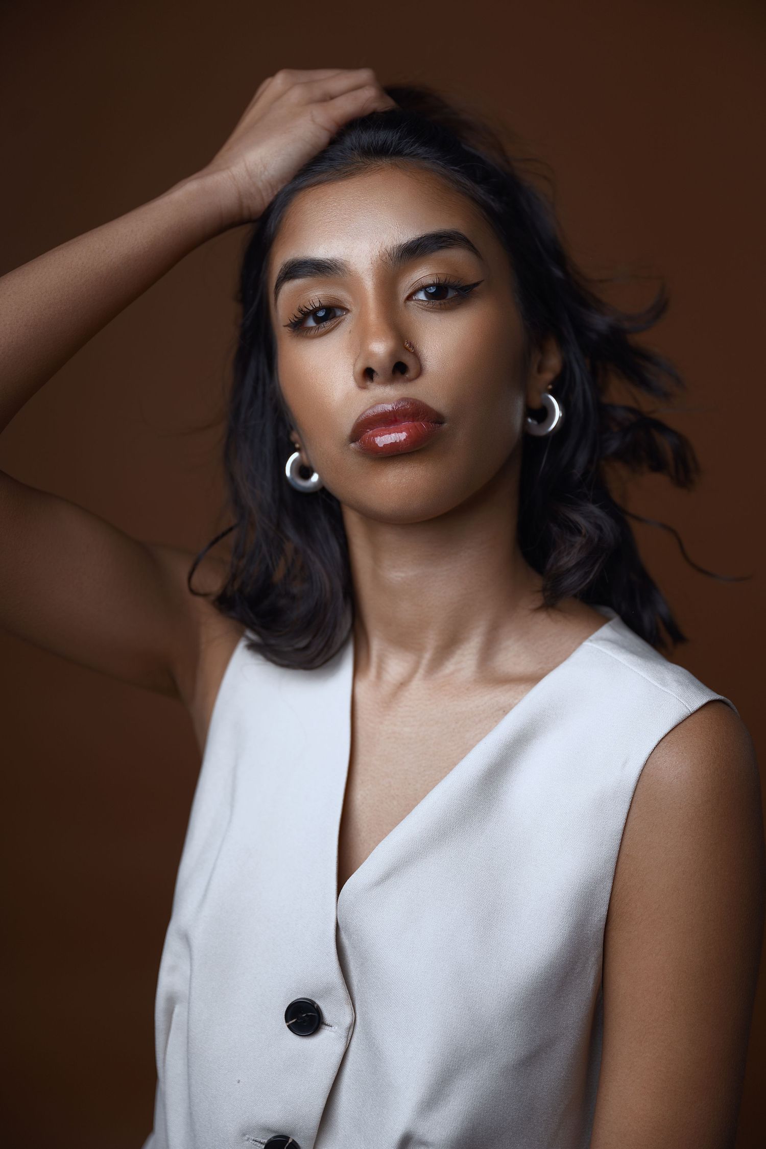 Fashion portrait with white sleeveless top and hoop earrings against brown background.
