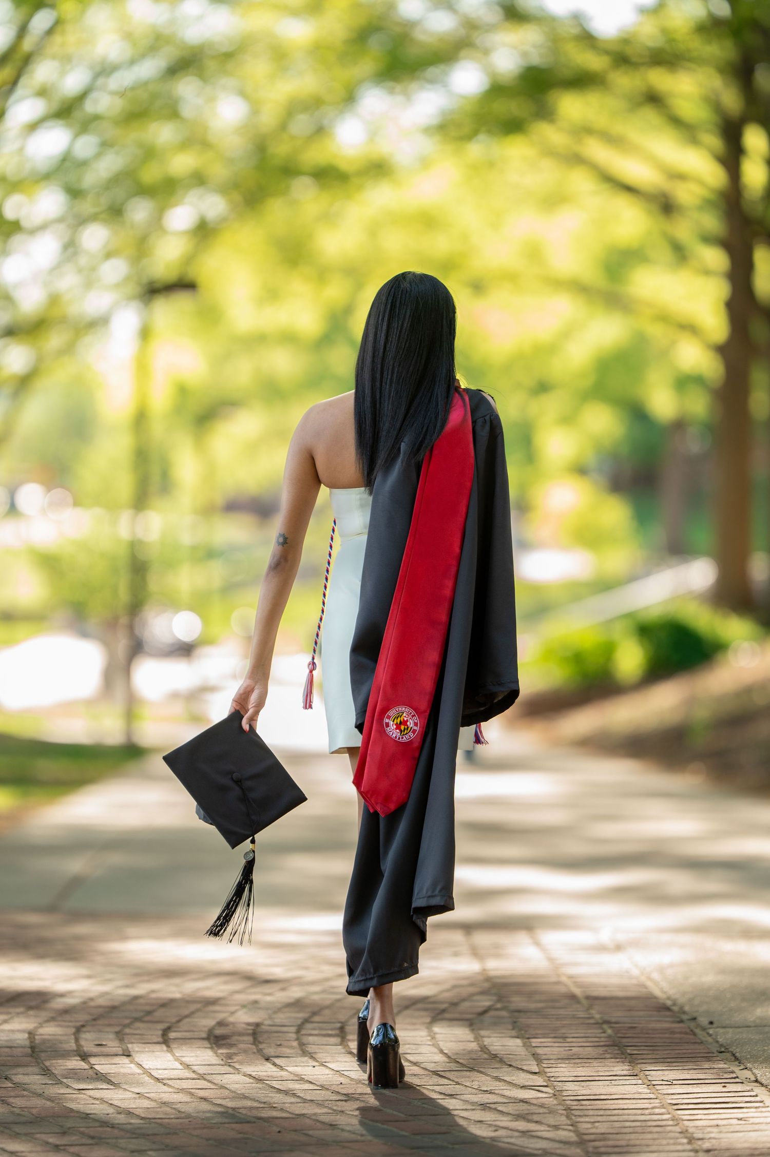 Graduate in cap and gown walking away on a curved path with trees in background.